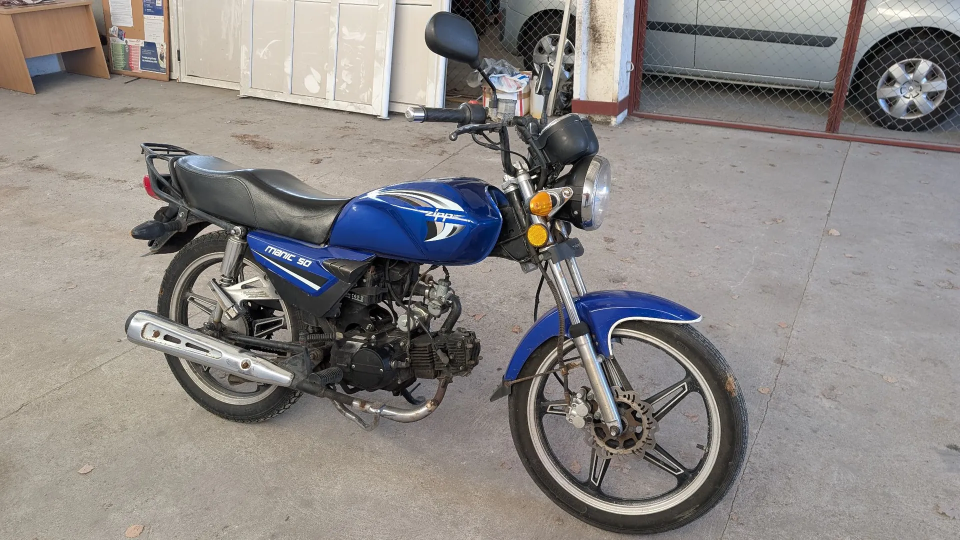 Niebieski motocykl marki Zipp, model Manic 50, zaparkowany na betonowym podłożu. Pojazd ma czarną kanapę, srebrny wydech oraz czarne felgi z pięcioma ramionami. Na baku widnieje napis 'Zipp', a na bocznej owiewce napis 'MANIC 50'. W tle widoczny jest fragment białego samochodu za metalową bramą oraz białe drzwi.