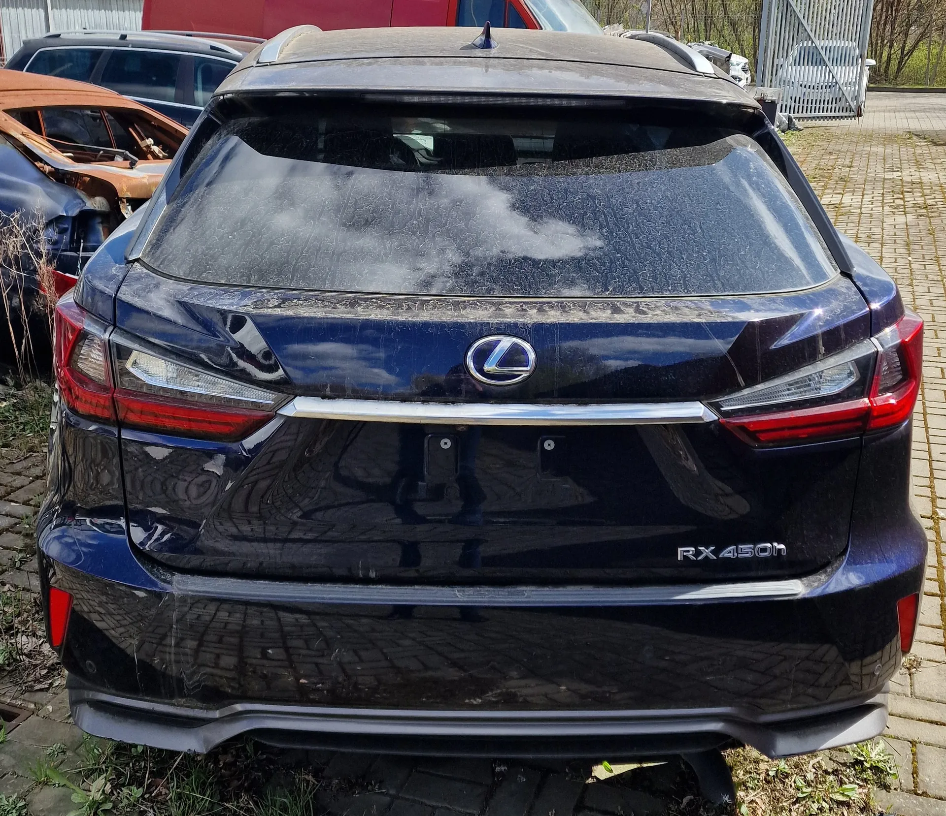 Tył ciemnoniebieskiego samochodu Lexus RX 450h z widocznym logo marki i napisem "RX 450h" po prawej stronie bagażnika. Przednia szyba bagażnika pokryta jest plamami lub kurzem. W tle widoczne są inne pojazdy, w tym uszkodzony pomarańczowy samochód oraz czerwony pojazd. Samochód stoi na brukowanej powierzchni, a w otoczeniu znajdują się trawa i metalowa brama.