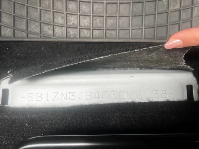 Zbliżenie na metalową tabliczkę z wygrawerowanym kodem ~SB1ZN3JE40E036138~. Tabliczka umieszczona jest na czarnym, miękkim podłożu. W prawym górnym rogu widoczny jest palec z różowym manicurem trzymający podarte czarne materiały. W tle siatkowata powierzchnia. Tekst na tabliczce jest czytelny i poziomy.