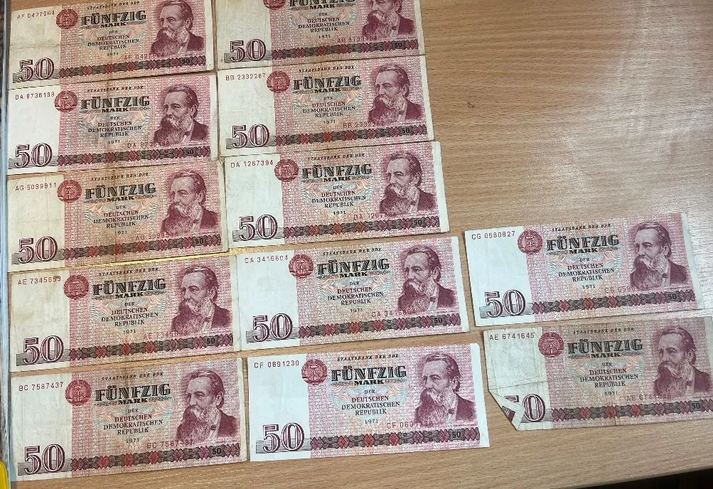 Kolekcja banknotów o nominale 50 marek z Niemieckiej Republiki Demokratycznej (NRD). Banknoty są ułożone w kilku rzędach na drewnianej powierzchni. Na każdym z nich widnieje portret mężczyzny w profilu oraz napis „FÜNFZIG MARK” i „DER DEUTSCHEN DEMOKRATISCHEN REPUBLIK”.