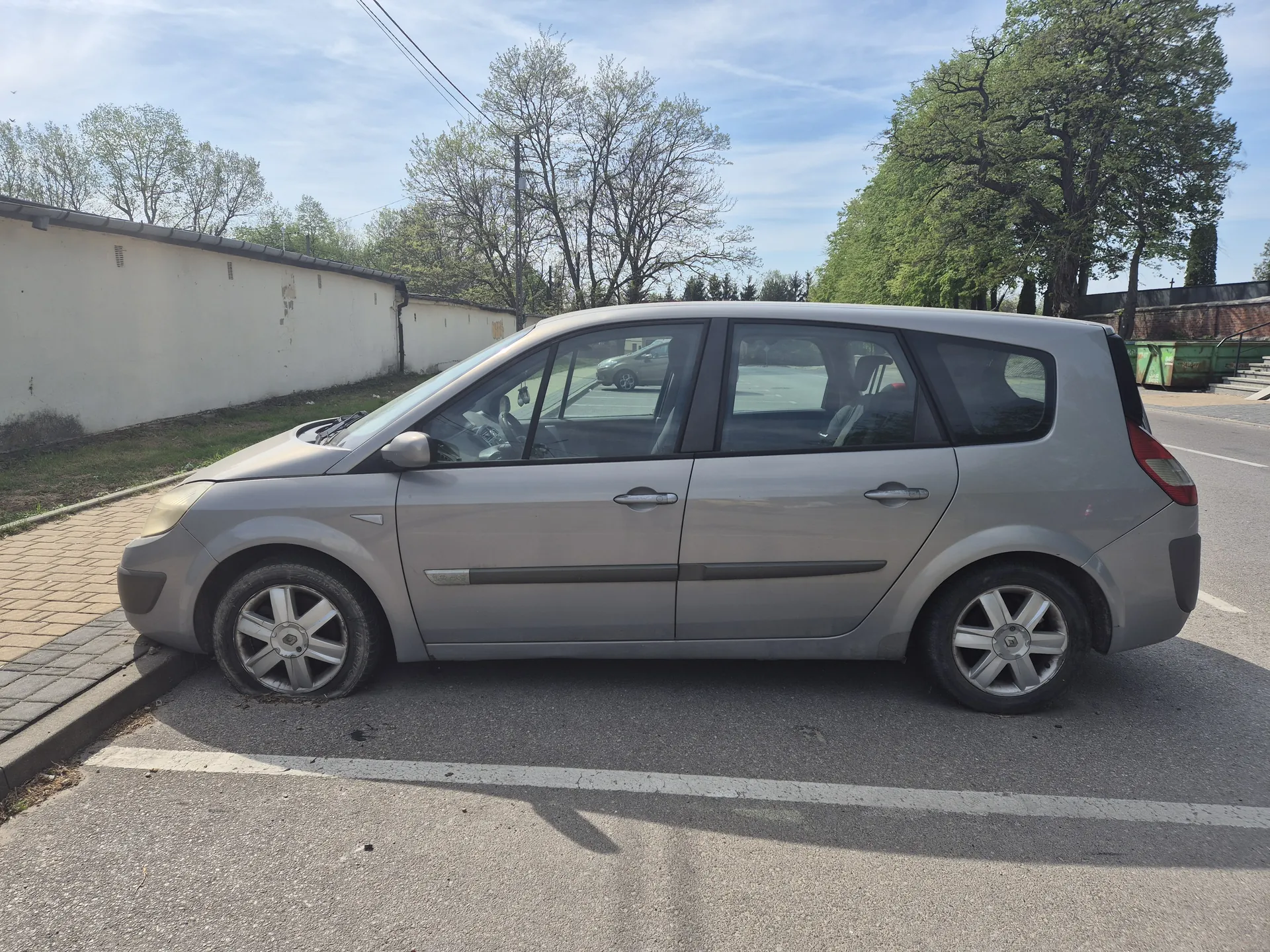 Srebrny samochód typu minivan zaparkowany na poboczu drogi. Po lewej stronie widoczna biała ściana, trawa i chodnik z kostki brukowej. W tle drzewa, niebo z chmurami oraz drugi samochód na parkingu. Droga z białą linią wyznaczającą pas ruchu.
