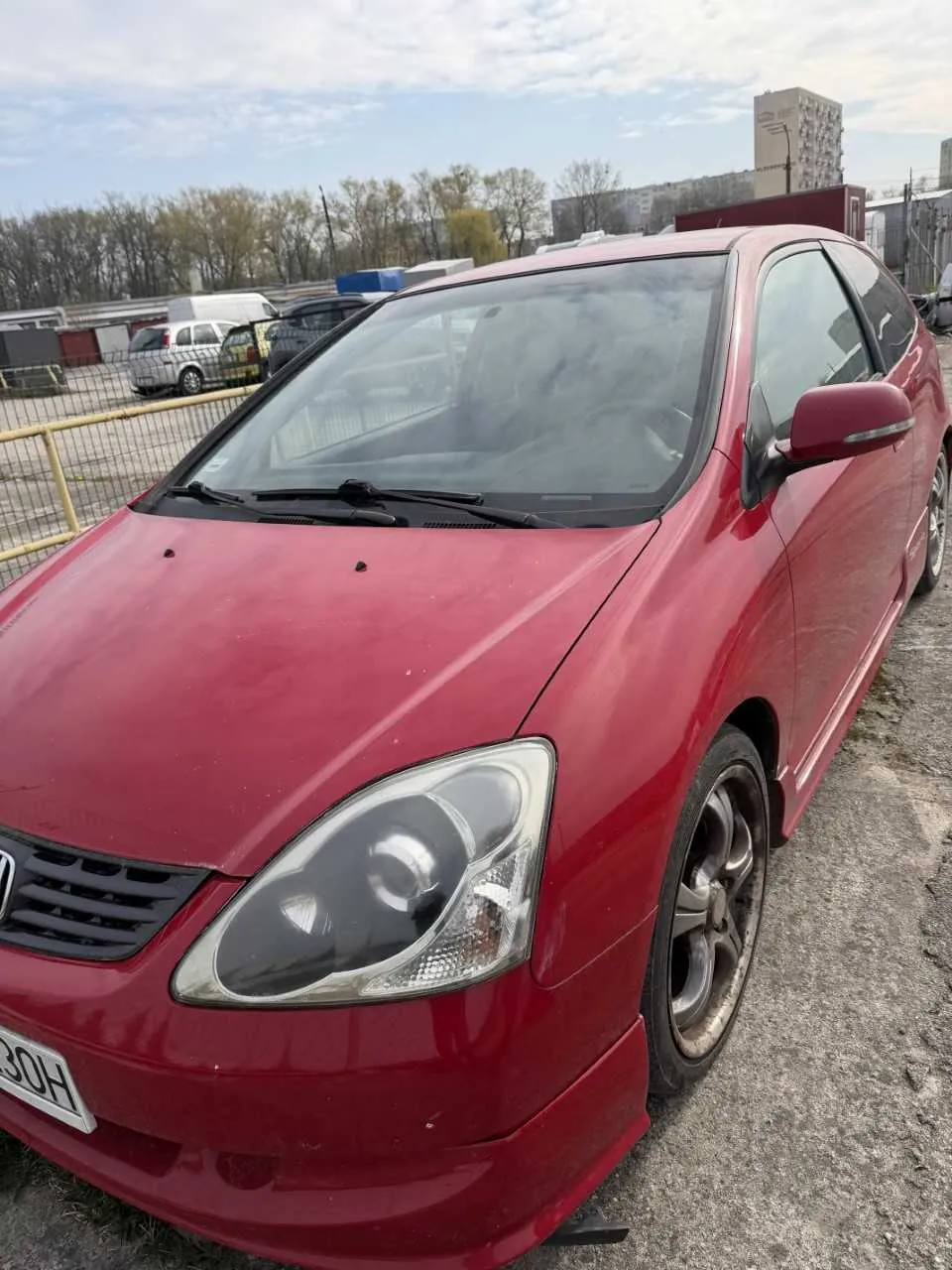 Czerwony samochód osobowy marki Honda z tablicą rejestracyjną 30H zaparkowany na otwartym placu. W tle widoczne są inne pojazdy, żółte ogrodzenie, drzewa oraz budynki. Niebo częściowo zachmurzone.