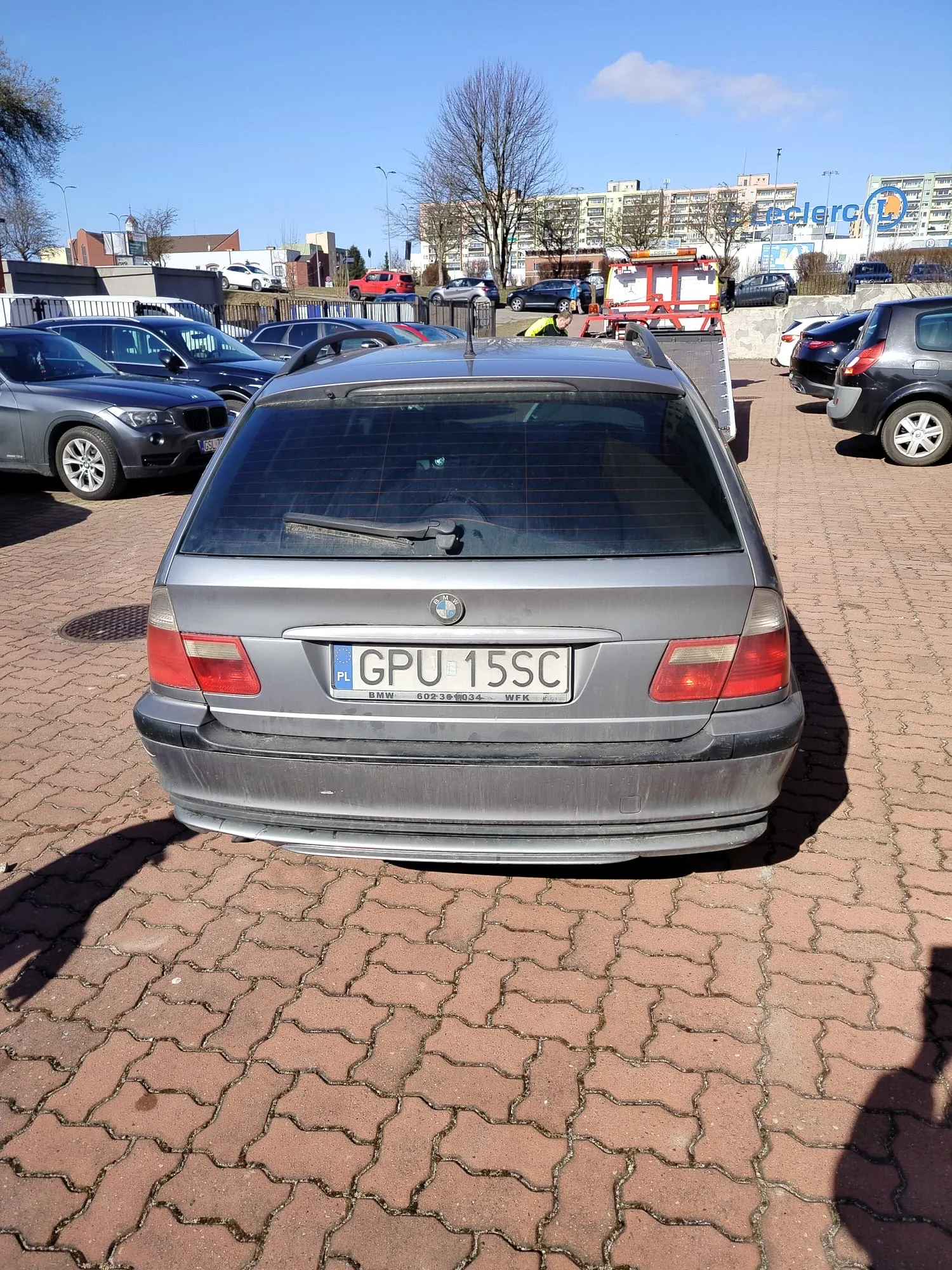 Z tyłu widoczny szary samochód marki BMW z tablicą rejestracyjną GPU 15SC (PL) na czerwono-białym tle. Samochód stoi na brukowanym parkingu, w tle inne zaparkowane pojazdy, budynki mieszkalne, drzewo bez liści oraz sklep Leclerc. Niebo jest jasne z kilkoma chmurami.