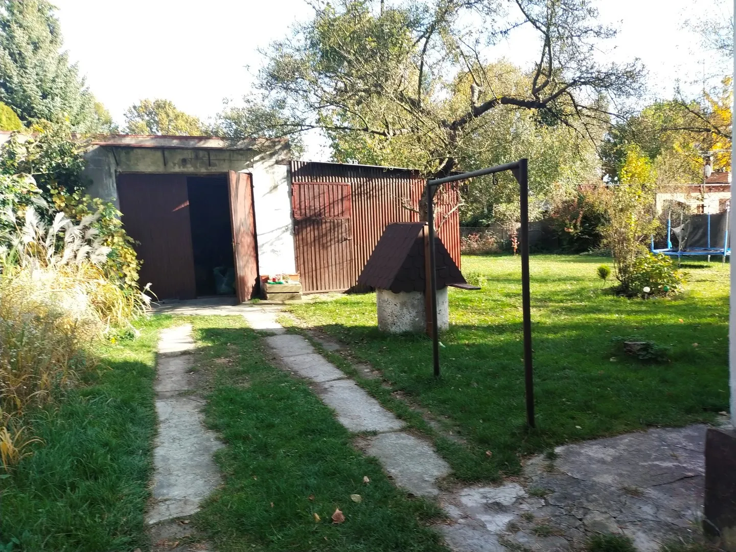 Zdjęcie przedstawia podwórze z trawnikiem i betonową ścieżką prowadzącą do garażu. Po lewej stronie znajduje się budynek z brązowymi, metalowymi drzwiami garażowymi. Obok garażu stoi drewniany płot oraz mała, biała konstrukcja przypominająca studnię z ciemnym, spiczastym daszkiem. Po prawej stronie widoczna jest metalowa rama oraz w oddali trampolina. W tle znajdują się liczne drzewa i krzewy pod jasnym niebem.