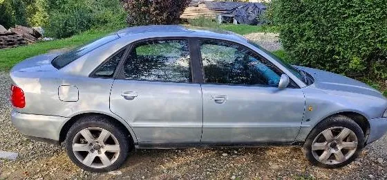 Srebrny samochód osobowy typu sedan zaparkowany bokiem na żwirowym podłożu. W tle widoczna jest zieleń krzewów i drzew oraz fragmenty zabudowań.