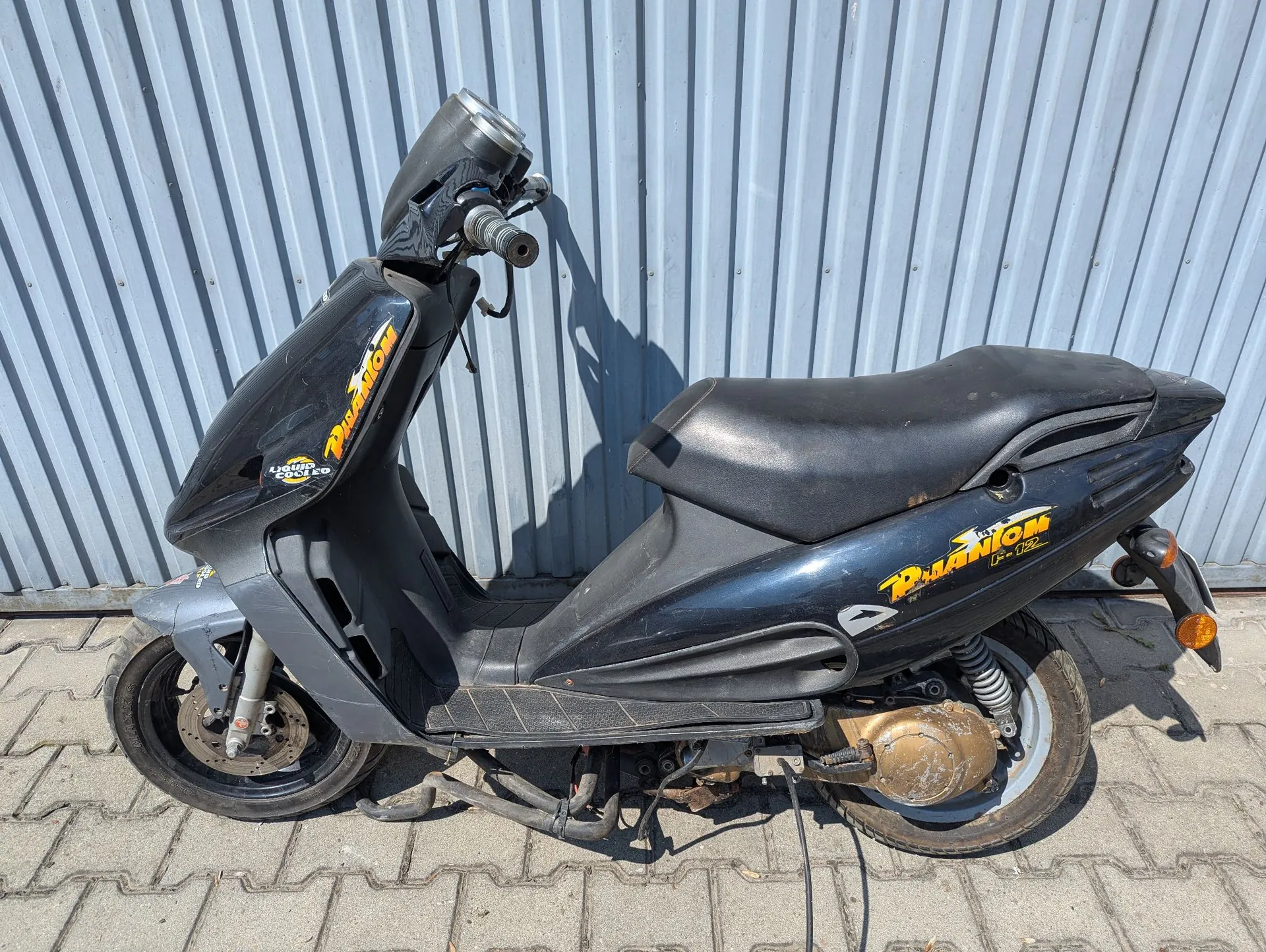 Czarny skuter zaparkowany bokiem na kostce brukowej na tle szarej, falistej blachy. Na boku skutera widnieje żółto-pomarańczowy napis 'RUANTON' oraz mniejszy napis '125'. Pojazd posiada czarne siedzenie, srebrne felgi i widoczny układ wydechowy u dołu.