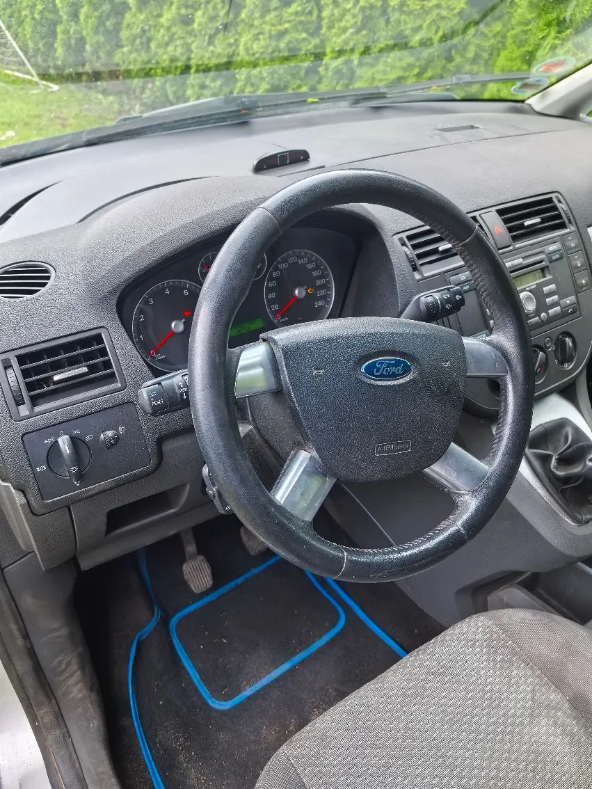 Wnętrze samochodu marki Ford z perspektywy kierowcy. Widoczna jest czarna kierownica z logo Ford, deska rozdzielcza z analogowymi wskaźnikami prędkości i obrotów, system audio oraz otwory nawiewów. Na podłodze znajduje się czarna mata z niebieskim obramowaniem.