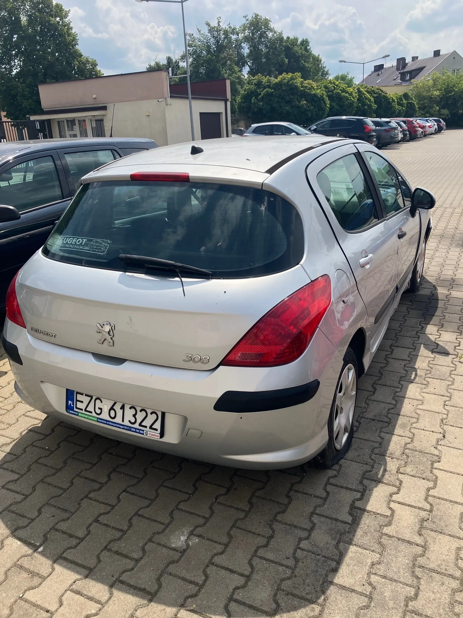 Srebrny samochód marki Peugeot 308 zaparkowany tyłem na kostce brukowej. Widoczna jest tylna część pojazdu z tablicą rejestracyjną EZG 61322. W tle znajdują się inne zaparkowane samochody, niski budynek oraz zielone drzewa pod jasnym, lekko zachmurzonym niebem.