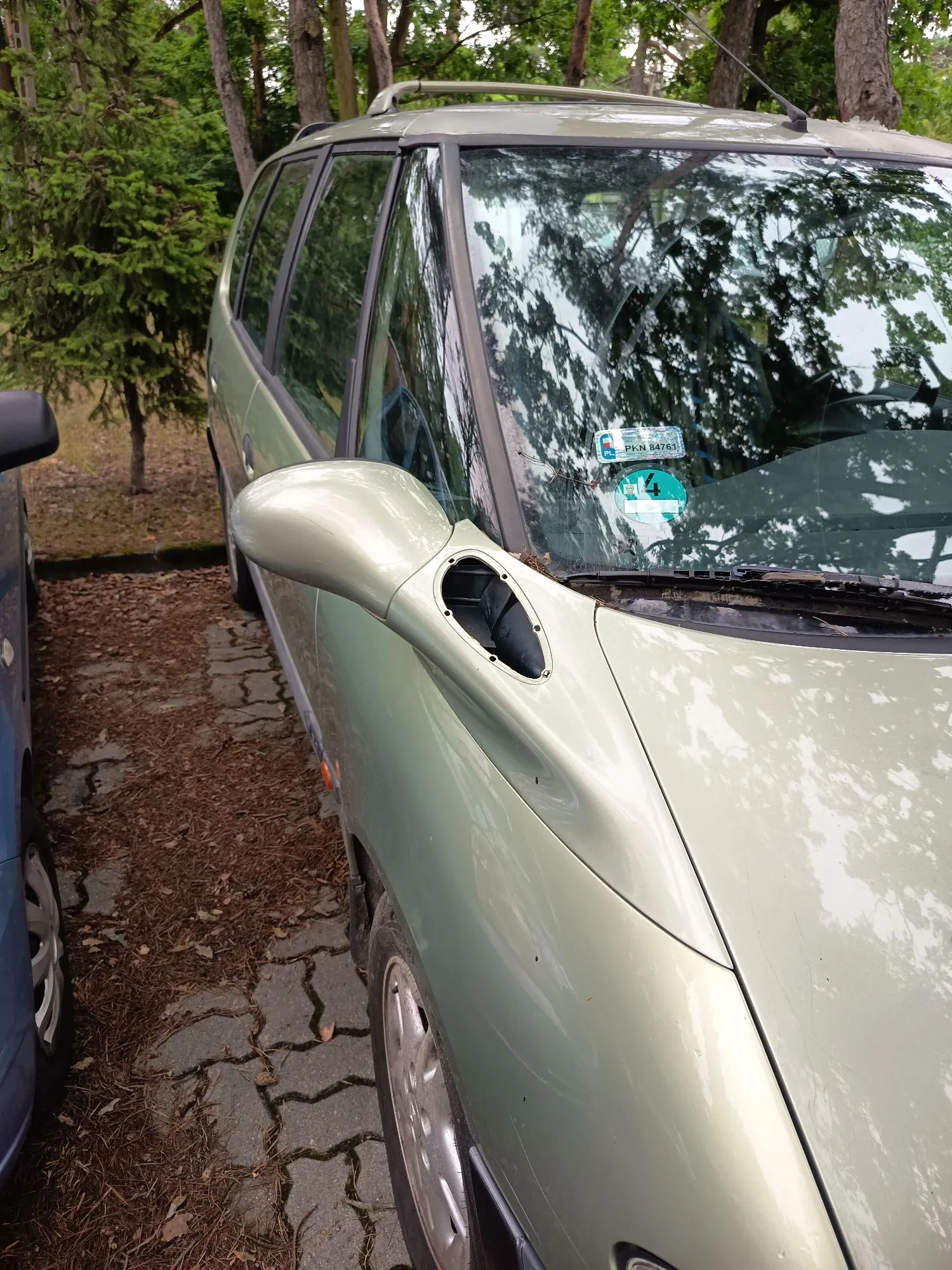 Zbliżenie na przednią prawą stronę jasnozielonego samochodu typu kombi zaparkowanego na kostce brukowej w otoczeniu drzew. Widoczna jest lusterko boczne, przednia szyba z naklejkami oraz fragment maski. Obok stoi fragment innego, niebieskiego samochodu.