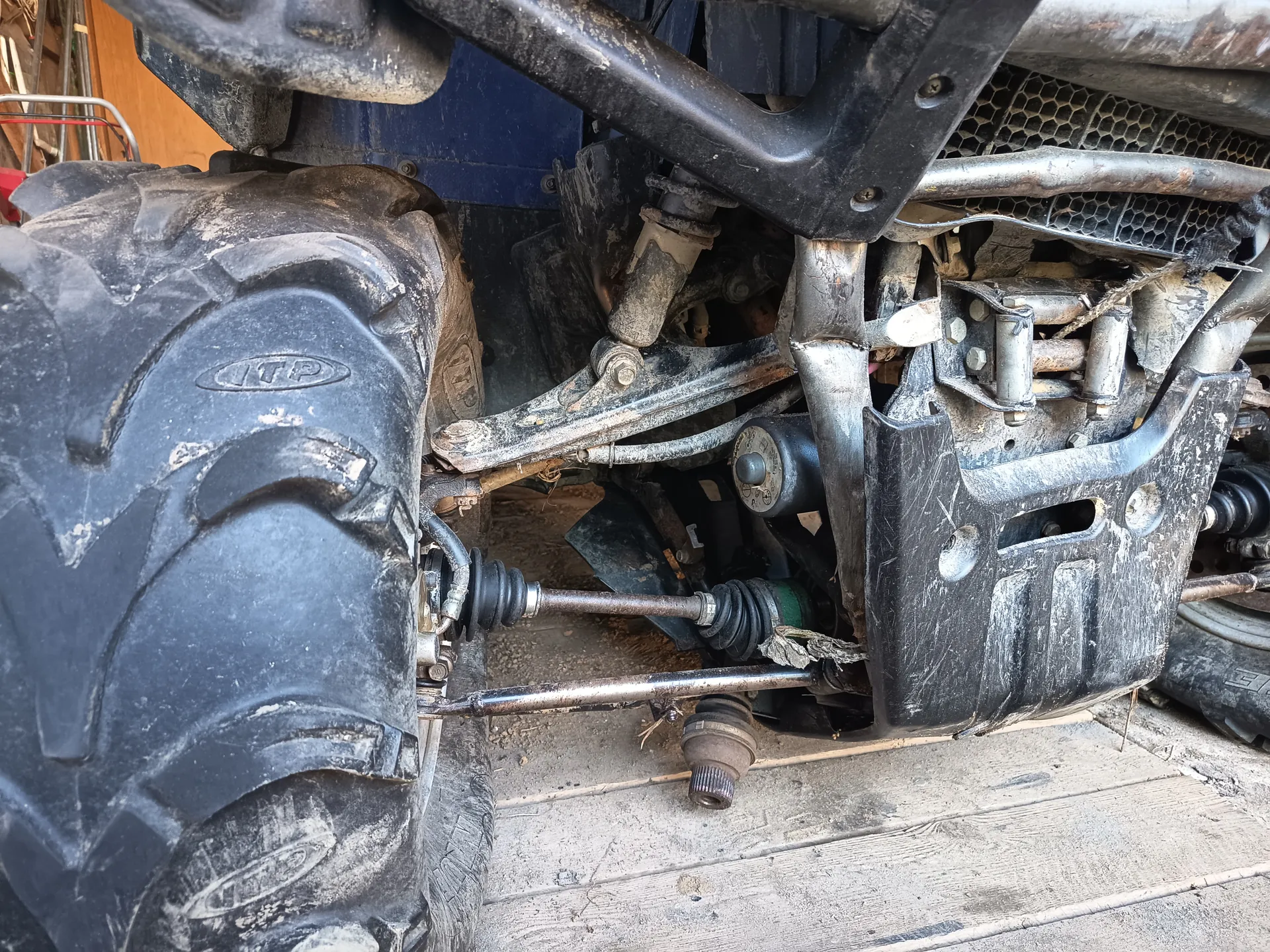 Zbliżenie na podwozie pojazdu, prawdopodobnie maszyny rolniczej lub terenowej. Widoczna duża, brudna opona z napisem 'ITP' po lewej stronie. Metalowe elementy podwozia, w tym zawieszenie, osie i przewody, pokryte kurzem i zabrudzeniami. W tle fragment niebieskiej części kadłuba oraz siatka chłodnicy. Podłoga z betonu z widocznymi śladami brudu i drewnianymi deskami.