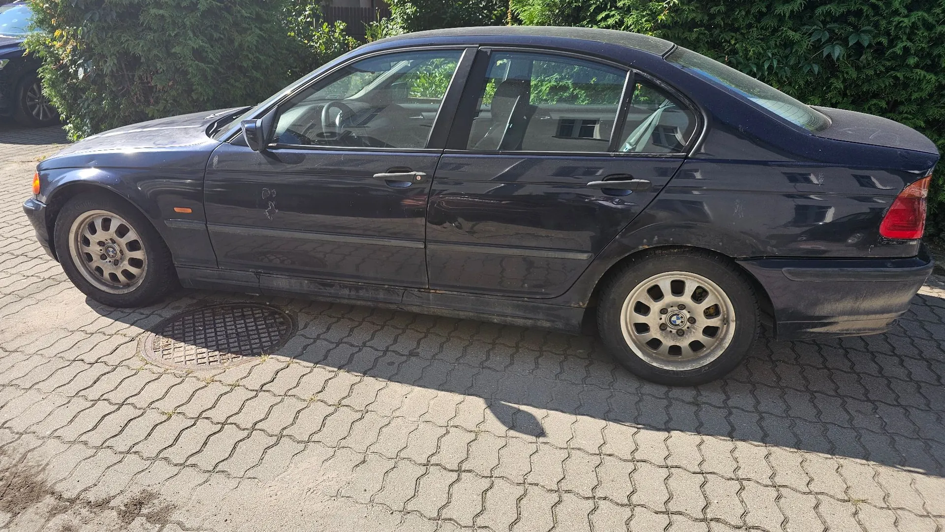 Ciemnoniebieski samochód osobowy marki BMW zaparkowany bokiem na kostce brukowej. Pojazd posiada widoczne uszkodzenia karoserii w postaci wgnieceń na drzwiach kierowcy oraz na tylnych drzwiach. W tle widoczne są zielone krzewy i drzewa.