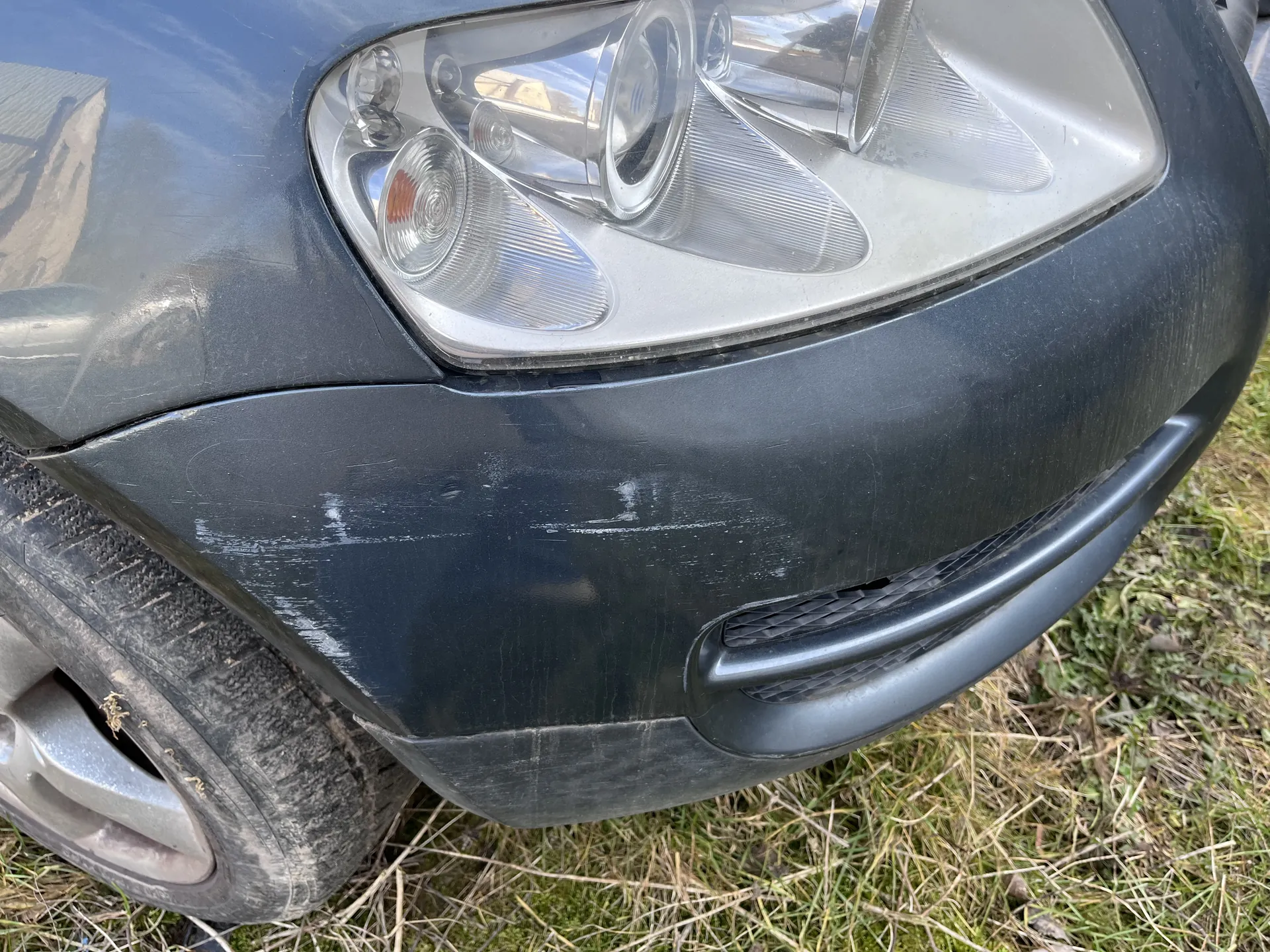 Zbliżenie przedniej części ciemnoszarego samochodu, w tym reflektora z pomarańczowym segmentem kierunkowskazu, widocznych zadrapań na zderzaku oraz części opony. Samochód stoi na trawie z zielonymi i suchymi pasmami.