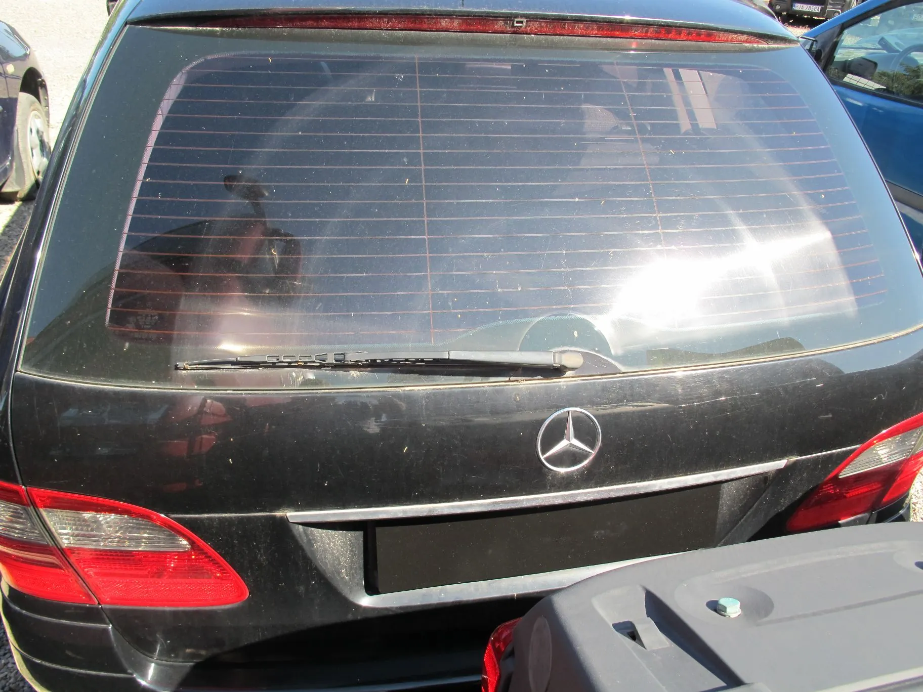 Tylny widok czarnego samochodu marki Mercedes-Benz. Widoczna jest tylna szyba z liniami ogrzewania, wycieraczka szyby oraz logo Mercedesa na klapie bagażnika. Poniżej logo znajduje się czarna, zakryta tablica rejestracyjna. Po bokach widoczne są czerwone lampy tylne. Samochód jest zakurzony.