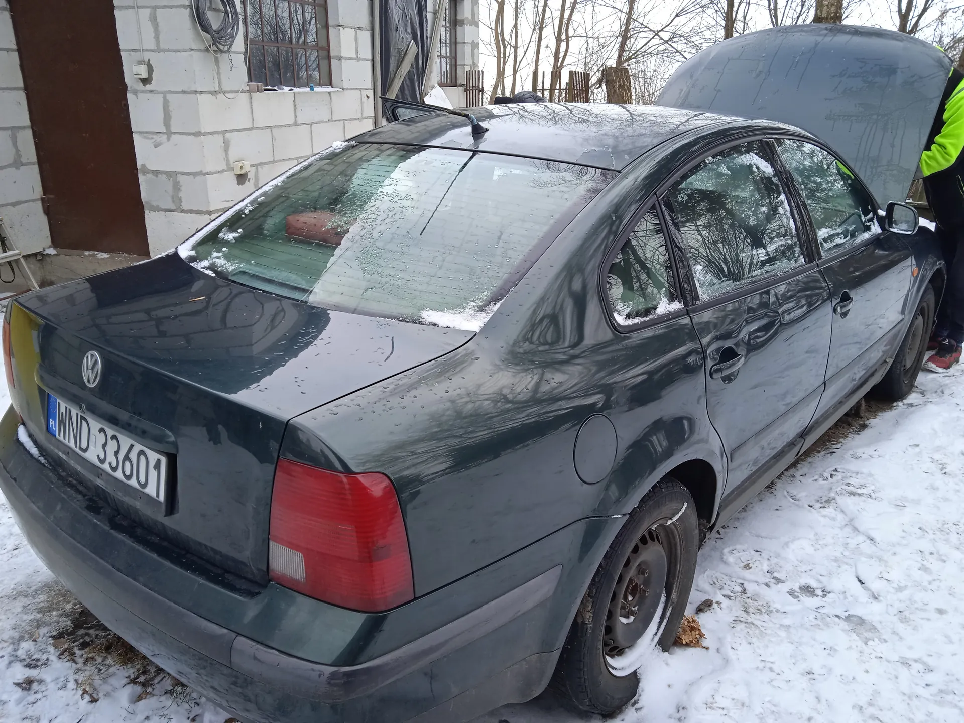 Ciemnozielony samochód osobowy marki Volkswagen z otwartą maską, zaparkowany na śnieżnej powierzchni. Widoczna tablica rejestracyjna WND 33601 z oznaczeniem PL. Po prawej stronie osoba w jasnozielonej kurtce odblaskowej. Tło: budynek z białych bloczków, brązowe drzwi, okna oraz nagie drzewa. Na dachu i szybach samochodu leży śnieg.