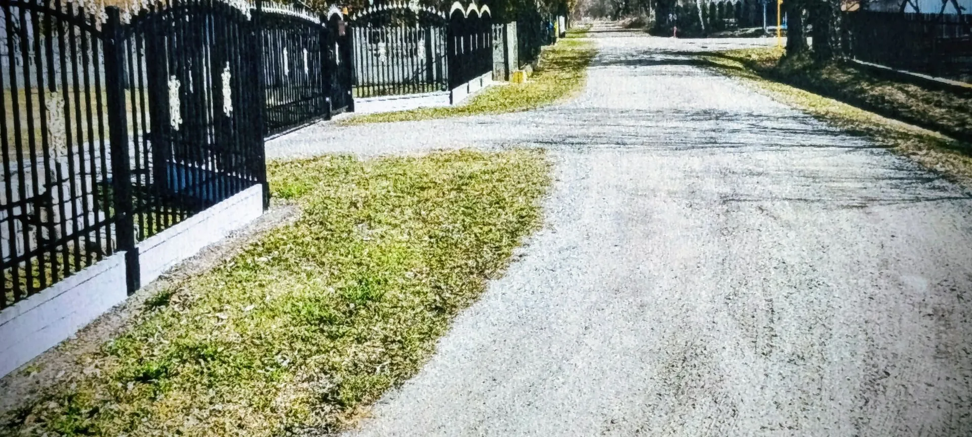 Widok na szutrową drogę prowadzącą w głąb obrazu. Po lewej stronie znajduje się czarny, metalowy płot z ozdobnymi elementami, osadzony na niskim, białym murku. Pomiędzy płotem a drogą widoczny jest pas trawy. W oddali droga kontynuuje swój bieg w stronę drzew i zabudowań.