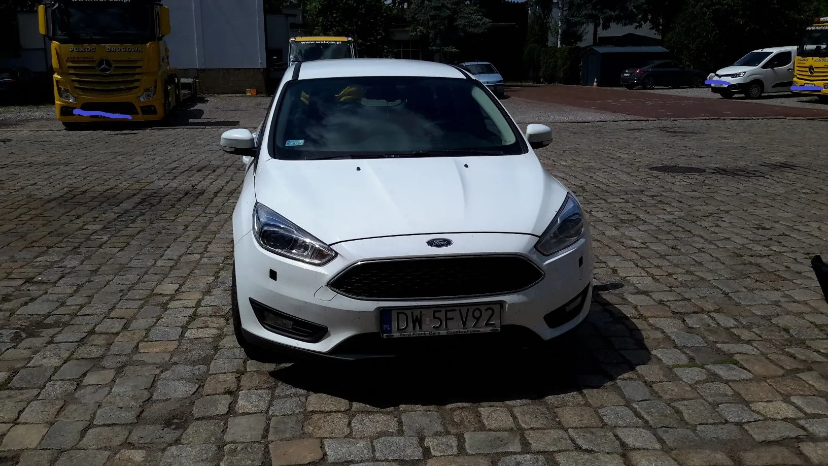 Biały samochód marki Ford zaparkowany przodem do widza na brukowanym placu. Pojazd posiada tablicę rejestracyjną o numerze DW 5FV92. W tle widoczne są inne pojazdy, w tym żółta ciężarówka z napisem 'POMOC DROGOWA ASSISTANCE' po lewej stronie oraz biały samochód dostawczy i kolejna żółta ciężarówka po prawej stronie.