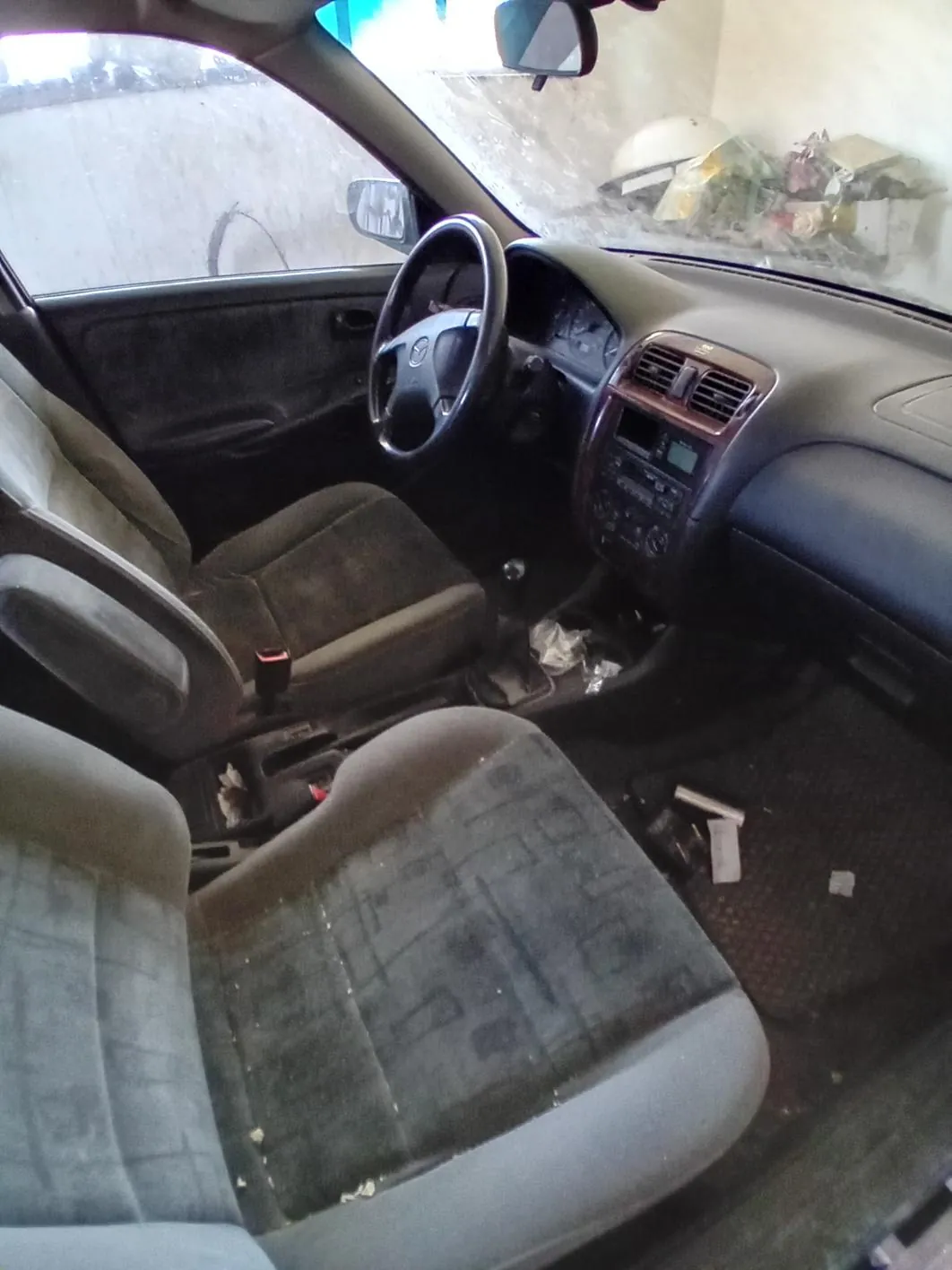 Wnętrze samochodu z brudnymi siedzeniami, widocznym kierownicą, deską rozdzielczą z odtwarzaczem i wentylacją. Na podłodze leżą śmieci i rozrzucone przedmioty. Przez przednią szybę widać zewnętrzne elementy, w tym fragment ściany i przedmioty poza pojazdem. Wnętrze wydaje się nieporządne i zapuszczone.