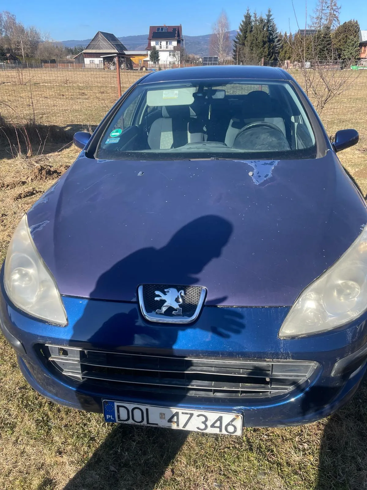 Niebieski samochód marki Peugeot z przodu, z widocznym logo lwa na atrapie chłodnicy. Tablica rejestracyjna PL DOL 47346. Cień osoby na masce. Tło: trawiaste pole, ogrodzenie, domy, drzewa i góry na horyzoncie. Jasne niebo. Wnętrze samochodu z widocznymi siedzeniami i kierownicą.