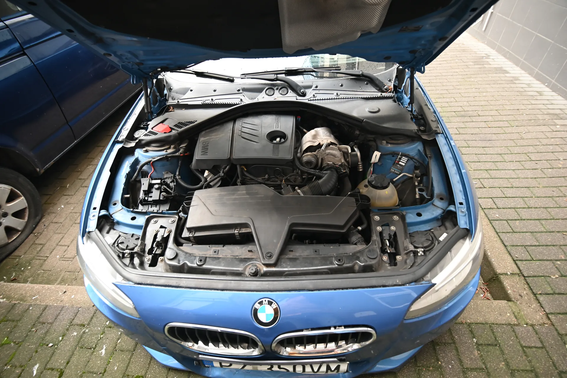 Niebieski samochód BMW z otwartą maską, ukazujący silnik z czarną pokrywą z napisem BMW, zbiornikiem płynu chłodniczego i innymi elementami. Tablica rejestracyjna P735UVM. Pojazd stoi na brukowanej nawierzchni obok ciemnoniebieskiego samochodu, przy szarej ścianie budynku.