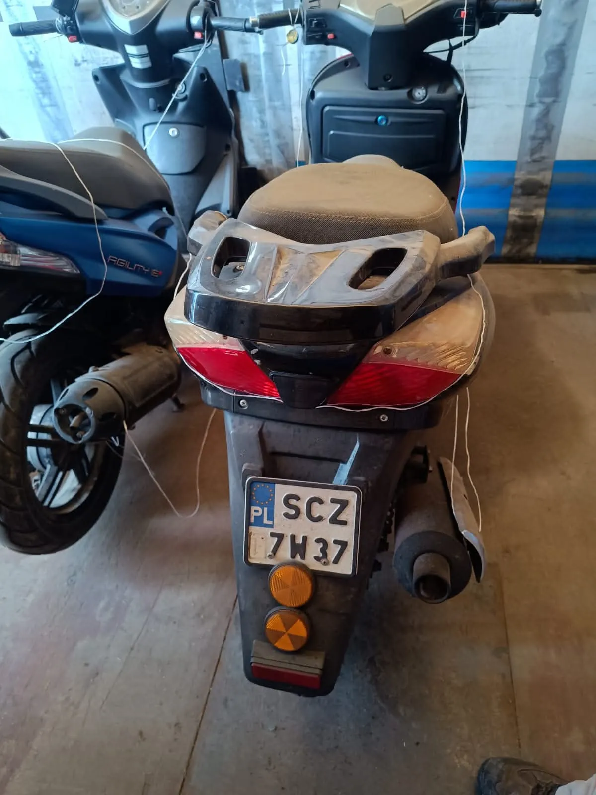 Tylny widok ciemnego skutera zaparkowanego w garażu obok innego, niebieskiego skutera. Widoczna jest tylna lampa, uchwyt dla pasażera oraz polska tablica rejestracyjna o numerze SCZ 7W37. Poniżej tablicy znajdują się dwa pomarańczowe odblaski.