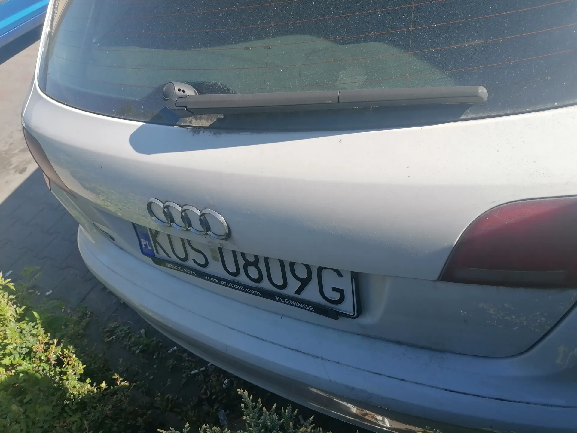 Zbliżenie na tylną część srebrnego samochodu marki Audi. Widoczne są cztery pierścienie logo Audi, tablica rejestracyjna z polskim oznaczeniem PL i numerem KUS 0809G. Na ramce tablicy rejestracyjnej znajduje się napis www.prutzbil.com oraz FLENINGE. W tle widać fragment chodnika, zielonych krzewów i częściowo niebieski obiekt. Samochód ma czarny wycieraczka tylnej szyby.