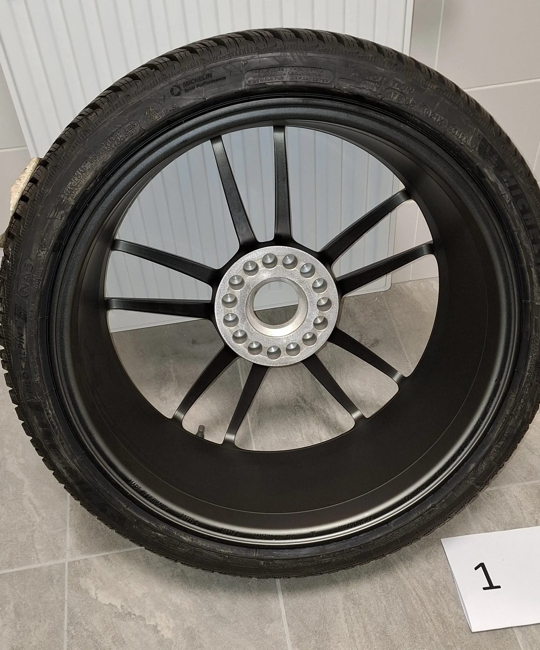 Czarna felga samochodowa z zamontowaną oponą marki Michelin, ustawiona pionowo na szarym kafelkowym podłożu. Felga ma wieloramienny wzór w kolorze matowej czerni i srebrny środek z otworami na śruby. W prawym dolnym rogu zdjęcia widoczna jest biała kartka z czarną cyfrą 1.