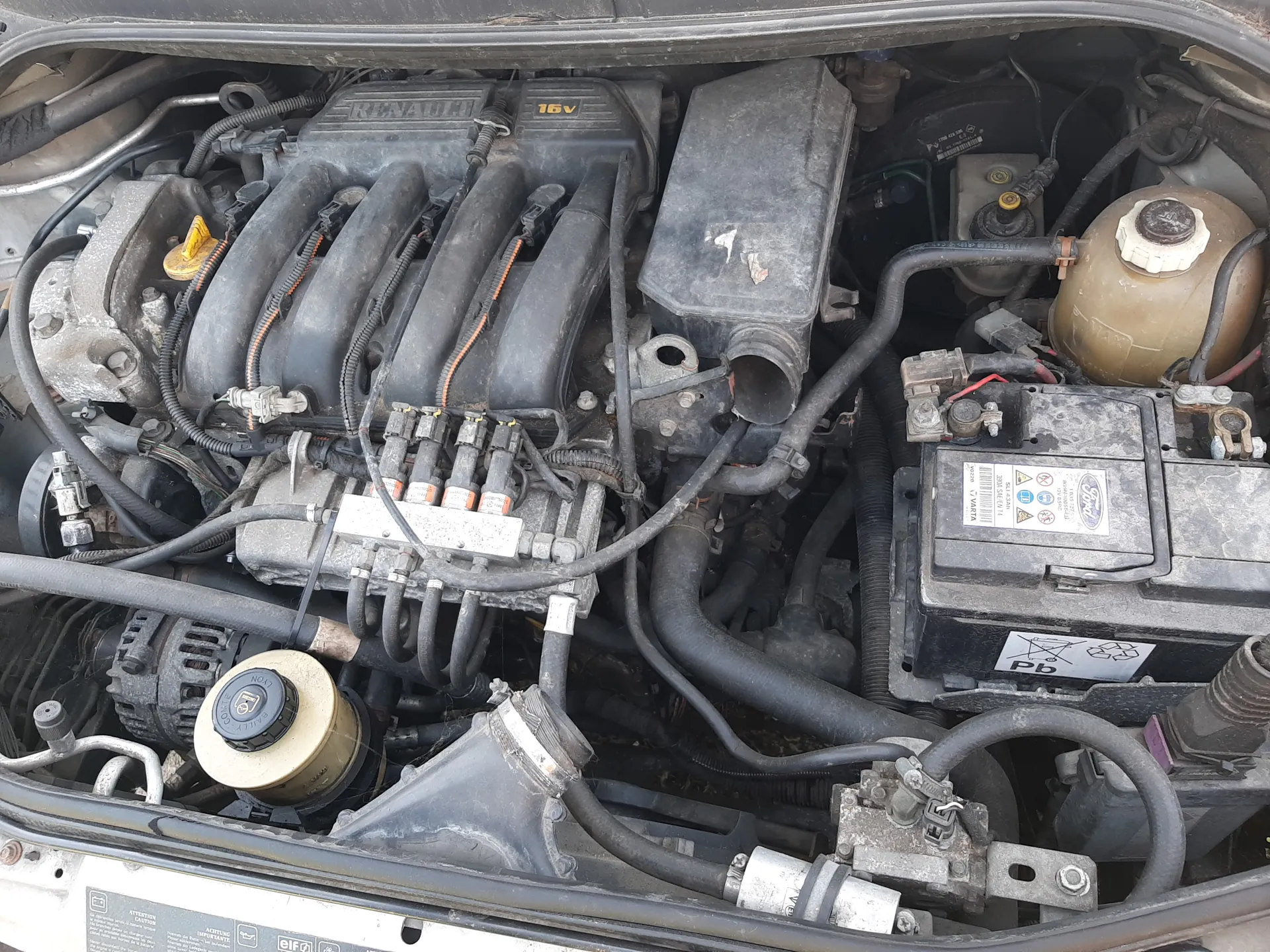 Silnik samochodu marki Renault z oznaczeniem 16V na pokrywie. Widoczne elementy: czarne przewody, żółty uchwyt miernika oleju, zbiornik chłodniczy, akumulator Ford z etykietą zawierającą symbole i napis Pb, zanieczyszczona powierzchnia silnika oraz różne przewody i rury. Na akumulatorze widoczne są również symbole ostrzegawcze i informacyjne.