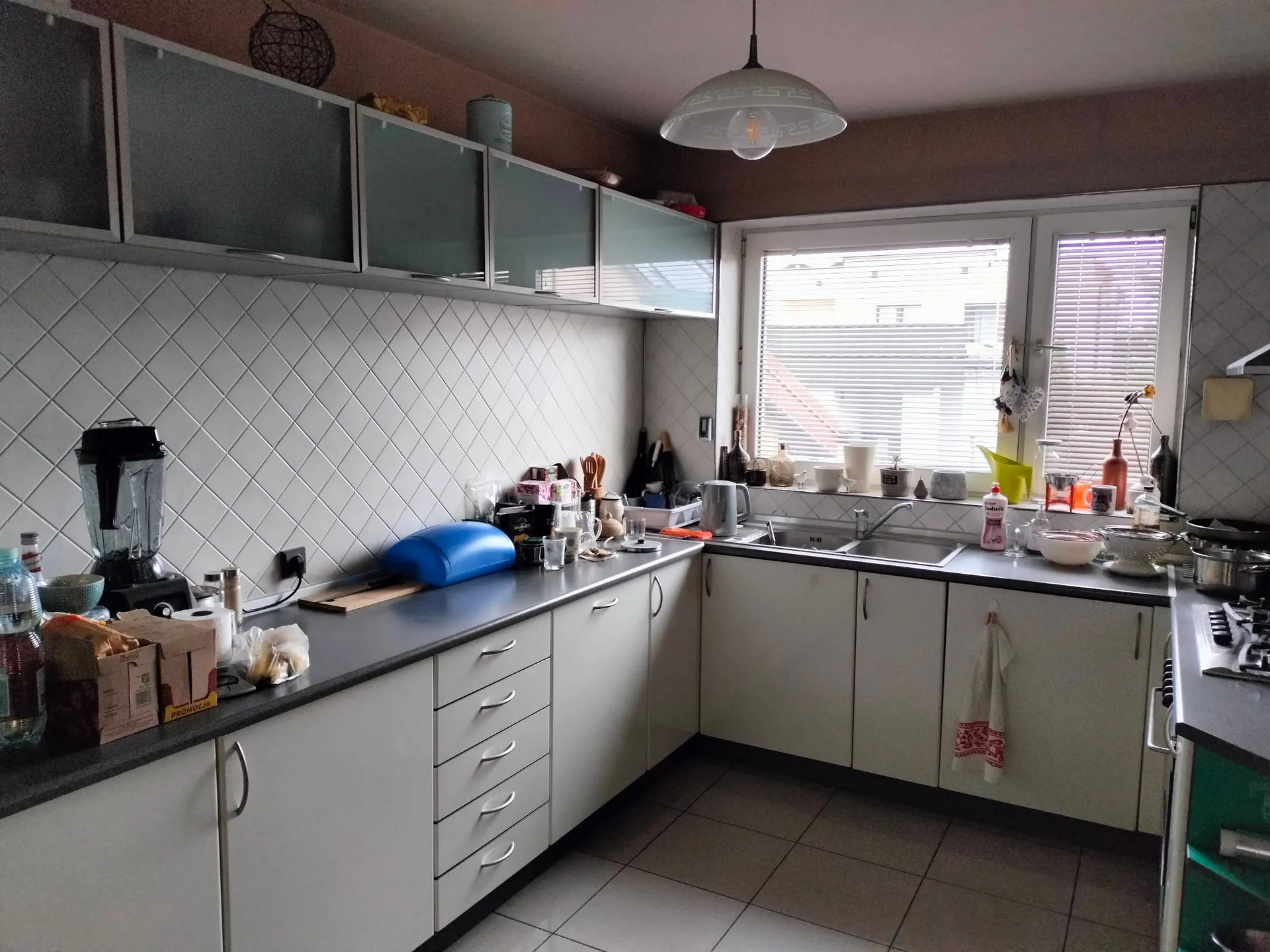 Wnętrze kuchni z białymi szafkami i szarym blatem. Na ścianie znajduje się biały kafel w romby. Na blacie ustawione są różne przedmioty: blender, niebieska mata do ćwiczeń, słoiki, przyprawy oraz naczynia. W tle widoczne jest okno z białymi ramami i żaluzjami, przez które wpada światło. Nad blatem zamontowane są szafki wiszące z matowymi, szklanymi frontami. Z sufitu zwisa biała lampa w kształcie kopuły.