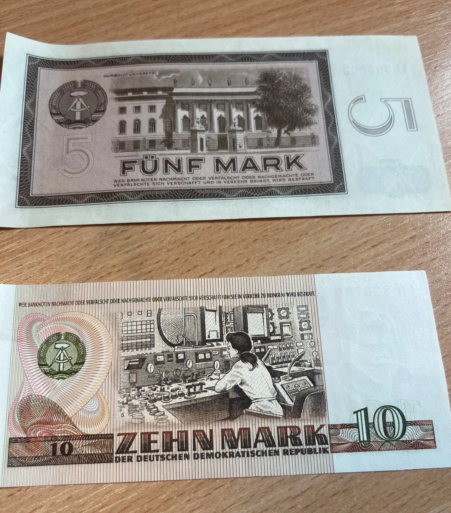 Dwa banknoty z Niemieckiej Republiki Demokratycznej (NRD) leżące na drewnianej powierzchni. Górny banknot o nominale 5 marek (FÜNF MARK) przedstawia budynek Uniwersytetu Humboldta. Dolny banknot o nominale 10 marek (ZEHN MARK DER DEUTSCHEN DEMOKRATISCHEN REPUBLIK) przedstawia kobietę pracującą przy panelu sterowania w centrum kontroli.