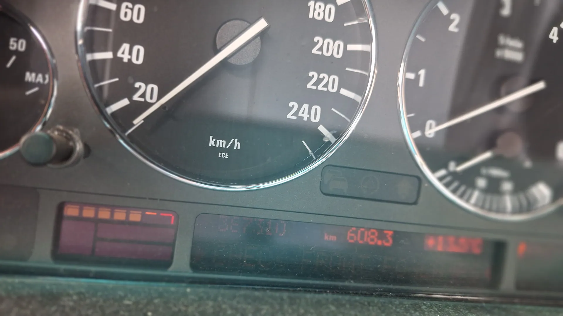Zbliżenie na deski rozdzielcze samochodu z trzema analogowymi wskaźnikami. Centralny wskaźnik prędkości oznaczony jako 'km/h' z zakresem do 240, wskazówka na ok. 20 km/h. Po lewej stronie częściowo widoczny inny wskaźnik z białymi cyframi i wskazówką. Po prawej stronie mały okrągły wskaźnik z napisem 'MAX' i cyfrą '50'. W górnej części wyświetlacze cyfrowe: czerwony wyświetlacz z tekstem '608.3 km' oraz nieczytelny wyświetlacz z cyframi. Widoczne również ikony ostrzegawcze i segmenty pomarańczowego wskaźnika po prawej stronie.