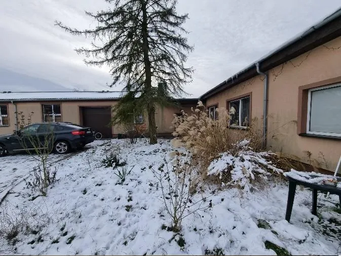 Zdjęcie przedstawia jednopiętrowy dom o beżowej elewacji z brązowymi obramowaniami okien i bramą garażową. Przed domem znajduje się zasneżony ogród z wysokim drzewem, suchymi krzewami i czarnym samochodem zaparkowanym po lewej stronie. Na prawo od drzewa widoczna jest czarna ławka. Dach domu pokryty jest śniegiem, a niebo jest zachmurzone. W tle widać poziomy horyzont i sąsiedni budynek.