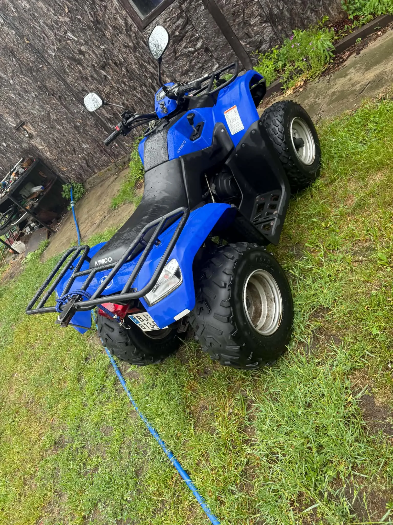 Niebieski quad marki Kymco zaparkowany na trawiastym terenie obok betonowej ścieżki i kamiennej ściany budynku. Pojazd posiada czarne opony z grubym bieżnikiem, czarne bagażniki z przodu i z tyłu oraz lusterka wsteczne. Na tylnej części widoczna jest tablica rejestracyjna o numerze BLN 813B.
