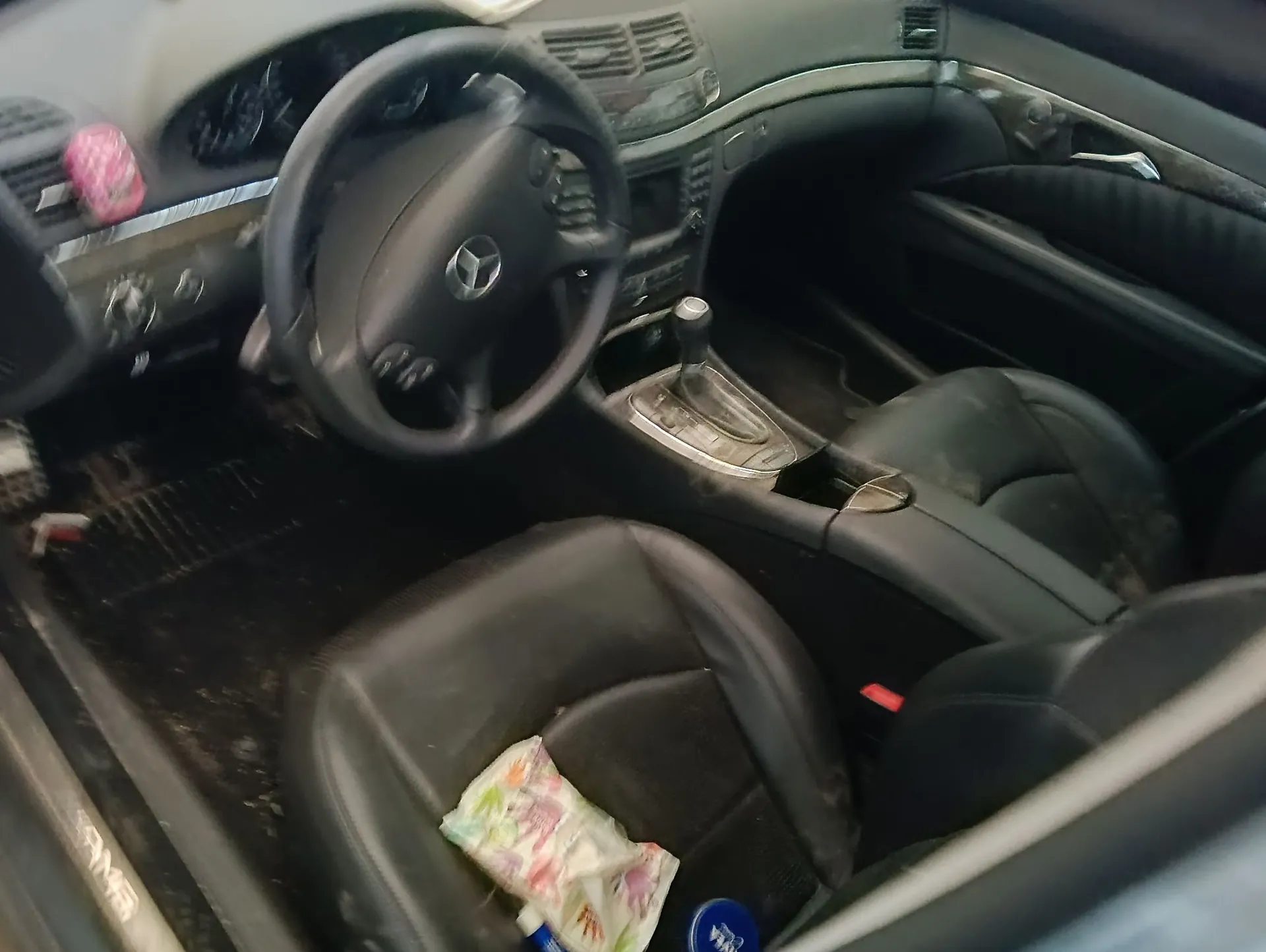 Wnętrze samochodu z widokiem na przednie fotele, kierownicę z logo Mercedes-Benz, deskę rozdzielczą oraz konsolę centralną z dźwignią zmiany biegów. Na fotelu pasażera leży kolorowa, plastikowa torebka. Wnętrze jest w kolorze czarnym i sprawia wrażenie zakurzonego.