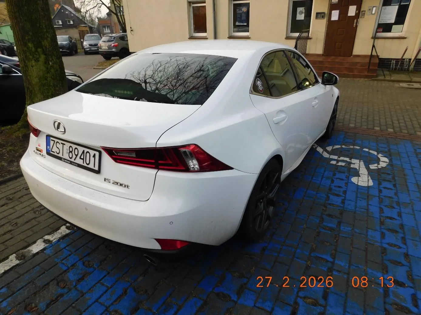 Biały samochód marki Lexus IS 200t zaparkowany na niebieskim miejscu parkingowym z białym symbolem niepełnosprawności. Tablica rejestracyjna ZST 89401. W tle budynek z brązowymi drzwiami, oknami i schodami, drzewo oraz inne zaparkowane pojazdy. Na dole obrazu widoczny znaczek daty i godziny: 27.2.2026 08:13. Podłoga z niebiesko-szarych płytek.