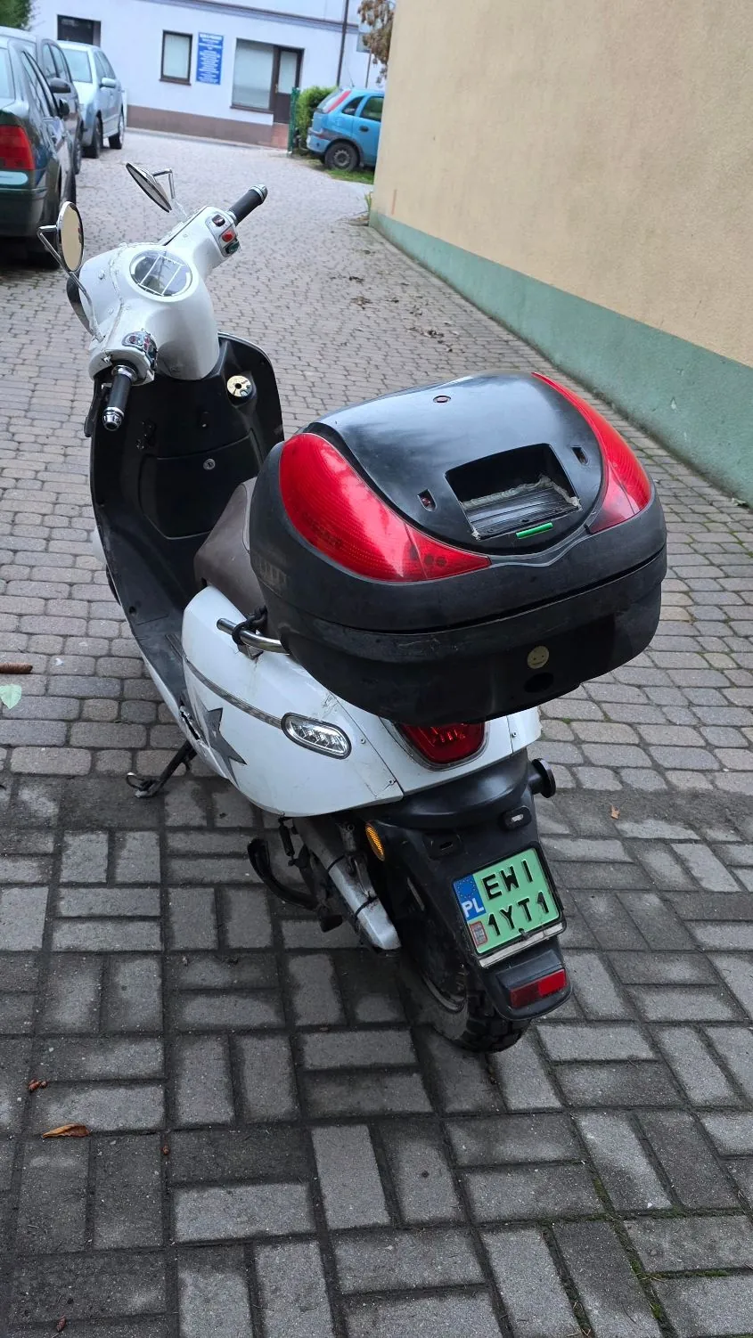 Biały skuter zaparkowany na kostce brukowej, widziany od tyłu. Skuter posiada czarny kufer z czerwonymi lampami z tyłu. Widoczna jest polska tablica rejestracyjna o numerze EWI 1YT1.
