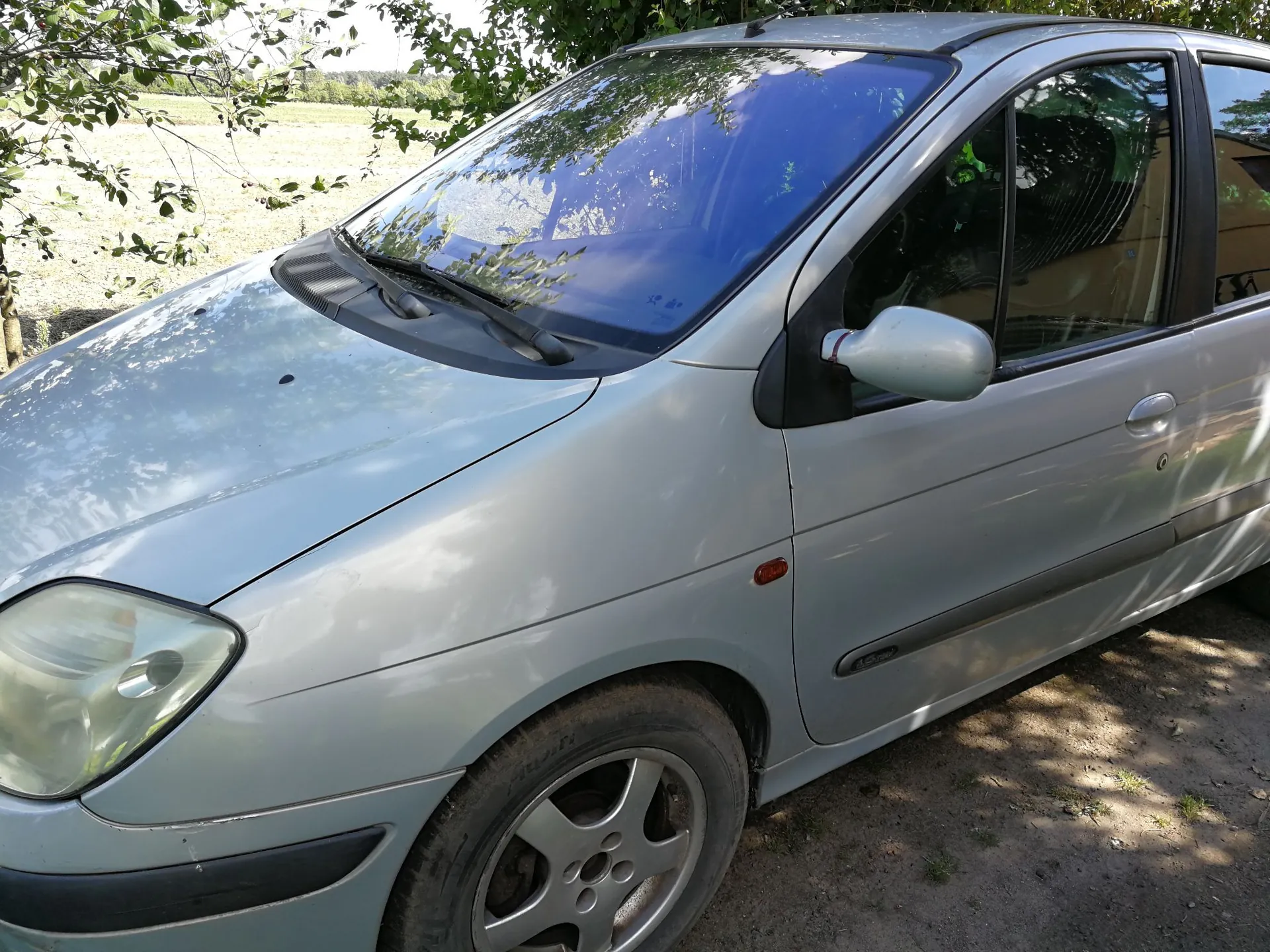 Srebrny samochód osobowy typu hatchback zaparkowany na piaszczystym podłożu. Widoczny jest przód i prawy bok pojazdu, w tym przednia lampa, maska, przednia szyba z niebieskim odcieniem oraz lusterko boczne. W tle widać zielone drzewa i jasne pole.