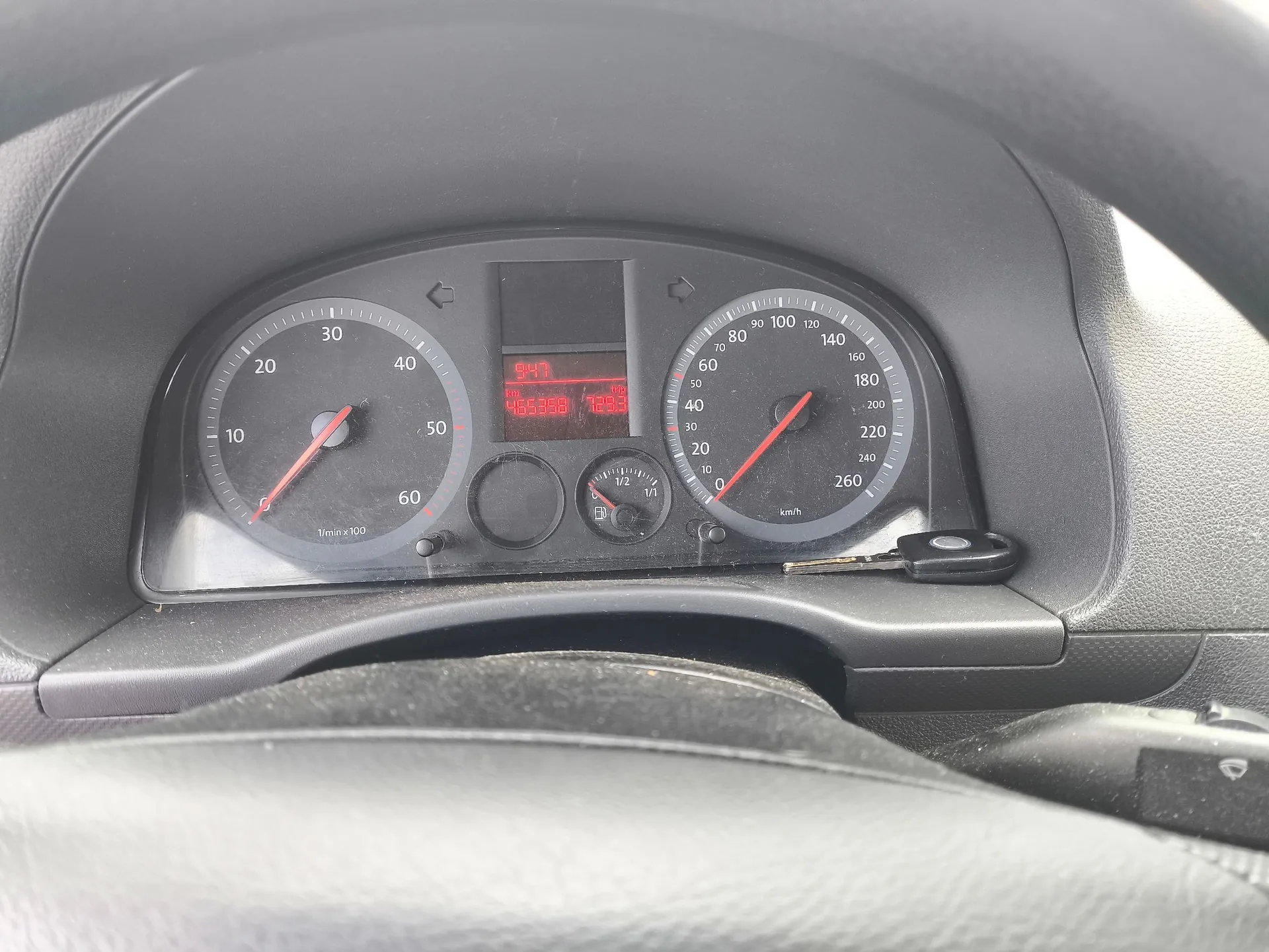 Zdjęcie z wnętrza samochodu przedstawiające panel wskaźników. Po lewej stronie znajduje się obrotomierz z zaznaczonymi wartościami od 10 do 60 (1/min x 100). Po prawej stronie znajduje się prędkościomierz z zaznaczonymi wartościami od 0 do 260 km/h. W centralnej części znajduje się cyfrowy wyświetlacz z czerwonymi cyframi wskazującymi godzinę 9:47, przebieg 485358 oraz temperaturę 12.3. Poniżej wyświetlacza widoczny jest wskaźnik poziomu paliwa. W prawym dolnym rogu panelu włożony jest kluczyk do stacyjki.