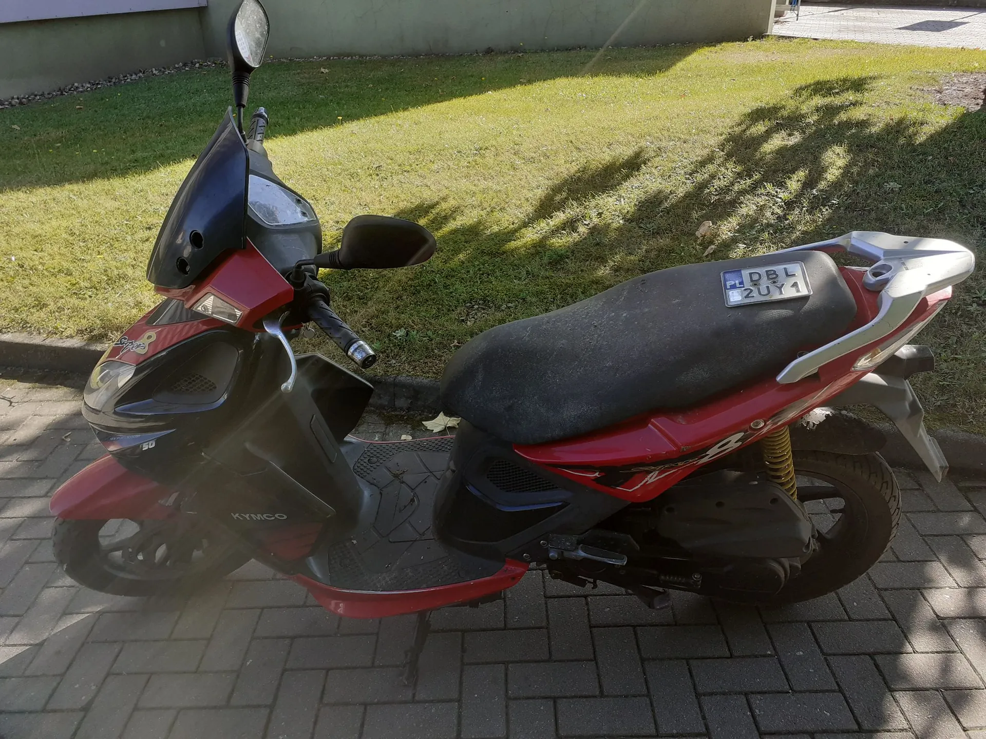 Czerwony skuter marki Kymco zaparkowany na kostce brukowej. Pojazd ma czarną kanapę, czarną owiewkę przednią i lusterka. Z tyłu widoczna jest tablica rejestracyjna o treści DBL 2UY1. W tle znajduje się trawnik i fragment budynku.