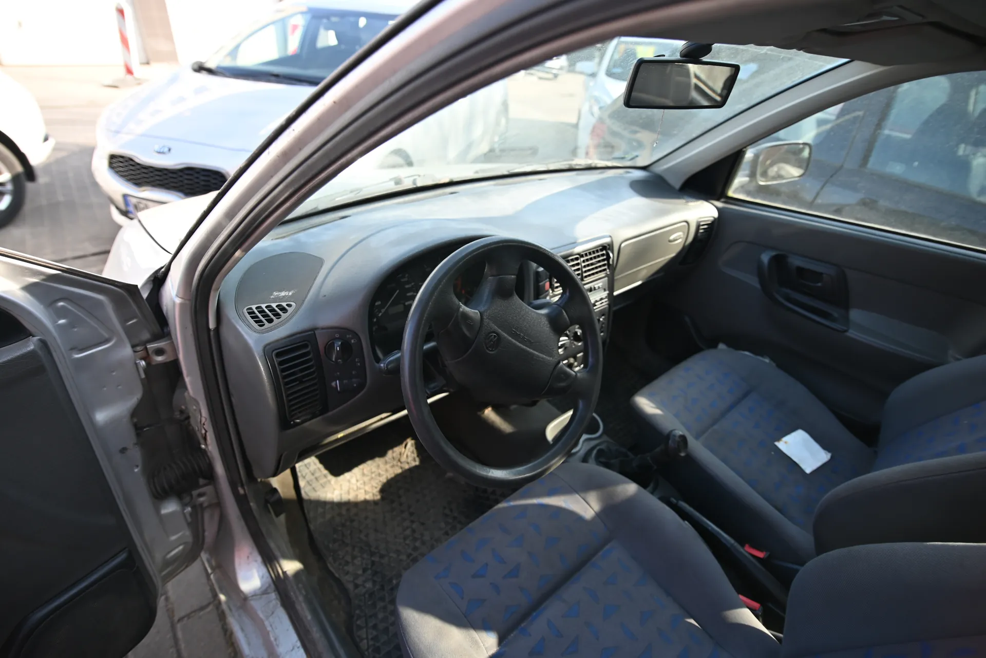 Wnętrze samochodu z otwartą drzwiami kierowcy. Widoczna kierownica z logo, deska rozdzielcza z wentylacjami i przyciskami, siedzenia z niebieskimi trójkątnymi wzorami. Na zewnątrz, na tle, srebrny samochód marki Kia z widocznym logo na atrapie chłodnicy oraz fragment białego pojazdu. Podłoga z kostki brukowej, w tle słup z czerwono-białą paską.