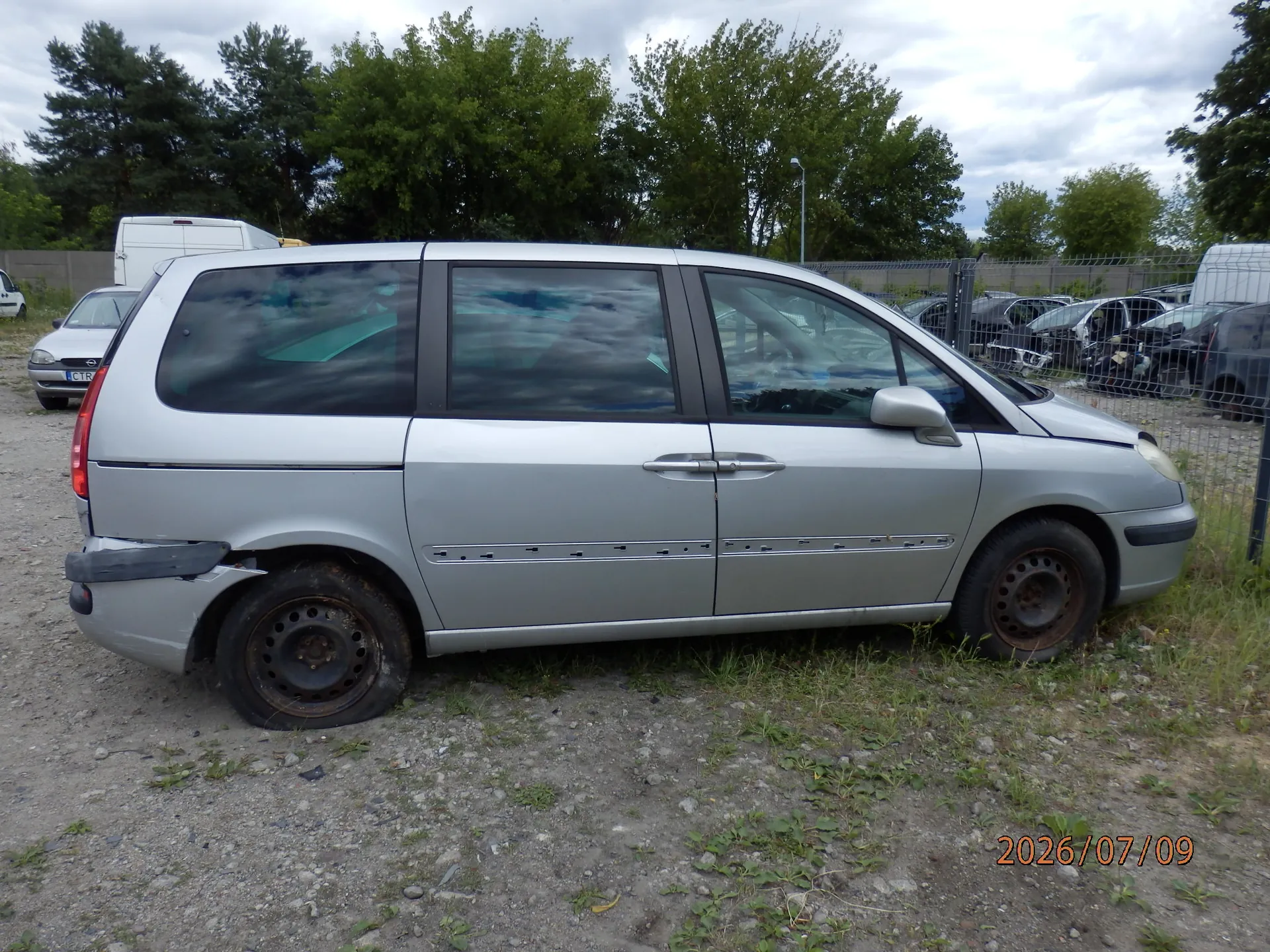 Srebrny samochód typu minivan zaparkowany bokiem na żwirowym podłożu. Pojazd jest uszkodzony w tylnej części nadkola oraz ma zniszczoną felgę tylnego koła. W tle widoczne są zielone drzewa, biały furgon oraz ogrodzenie z siatki, za którym stoją inne samochody. W prawym dolnym rogu zdjęcia znajduje się data 2026/07/09.