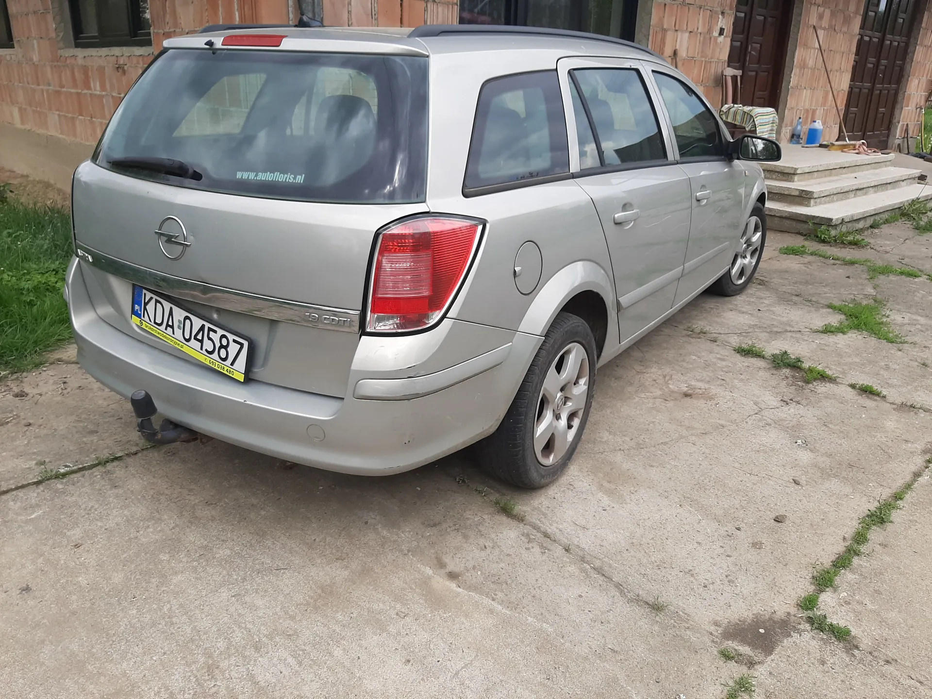 Srebrny samochód osobowy typu kombi marki Opel zaparkowany na betonowej powierzchni. Widok od tyłu z lewej strony. Na szybie tylnej widoczny napis www.autofloris.nl. Tablica rejestracyjna z numerem KDA 04587 z żółtą listwą na dole. W tle ceglany budynek z brązowymi drzwiami i schodami prowadzącymi do wejścia. Na ziemi rośnie trawa w szczelinach betonu.