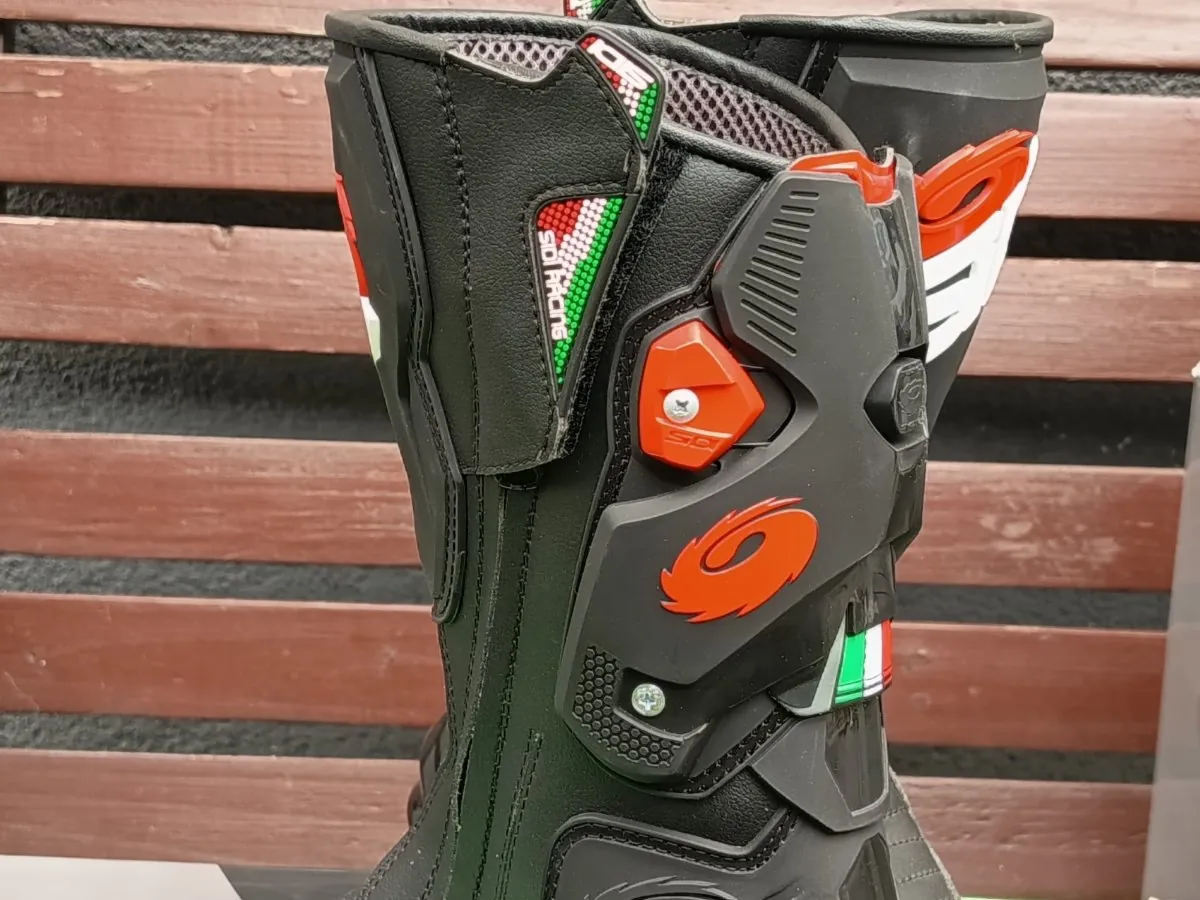 Zbliżenie na czarny but motocyklowy z czerwonymi i białymi akcentami. But posiada plastikowe wzmocnienia, klamry oraz ozdobny pasek w kolorach flagi włoskiej (zielony, biały, czerwony). Na boku widoczne jest czerwone logo w kształcie spirali oraz napis 'SIDI'. Tło stanowi brązowa drewniana konstrukcja z poziomych listew.