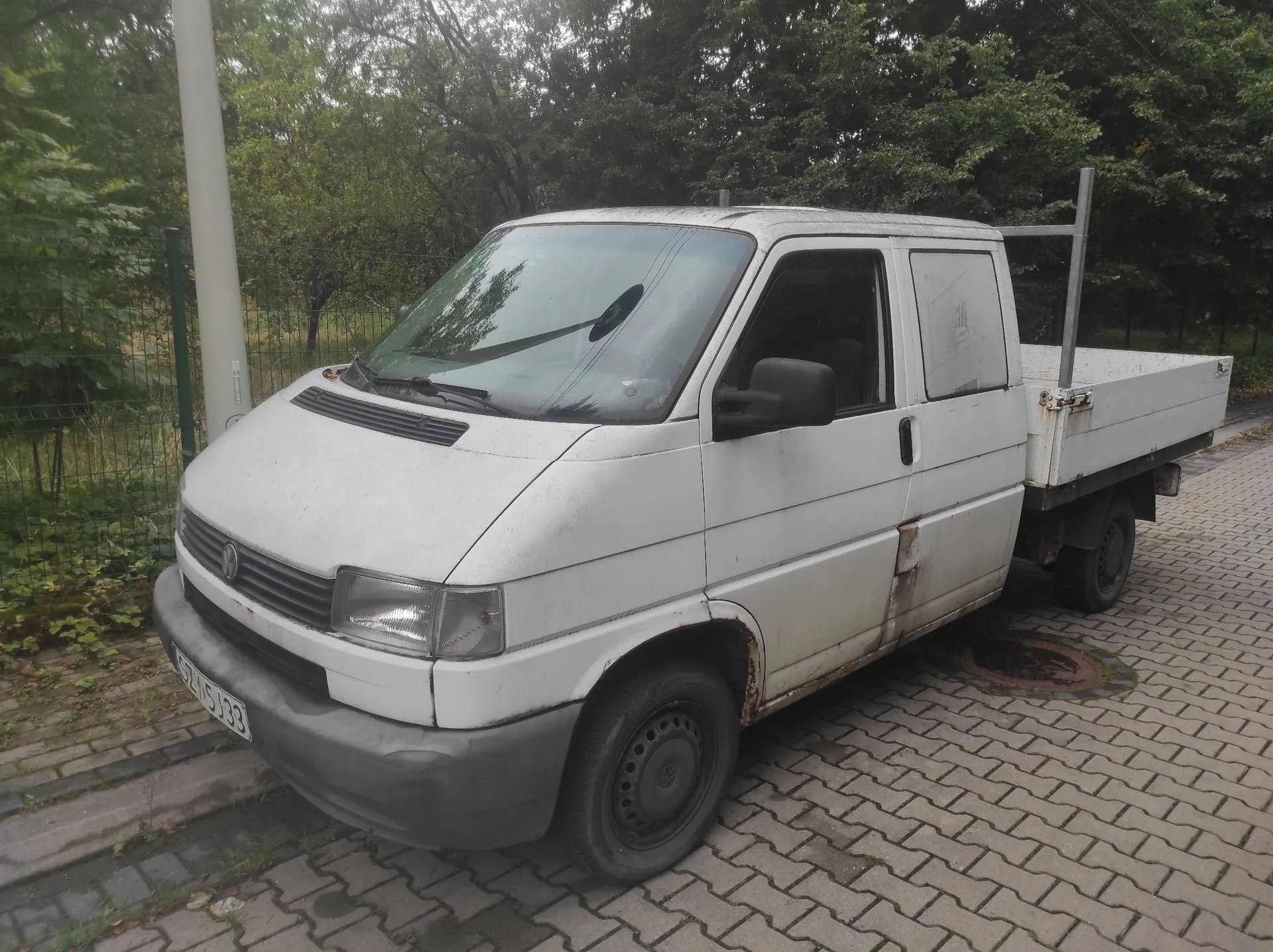 Biały samochód dostawczy typu pick-up zaparkowany na kostce brukowej. Pojazd jest brudny, z widocznymi śladami rdzy na dolnych partiach nadwozia. W tle znajdują się gęste, zielone drzewa i krzewy oraz biały słup oświetleniowy.