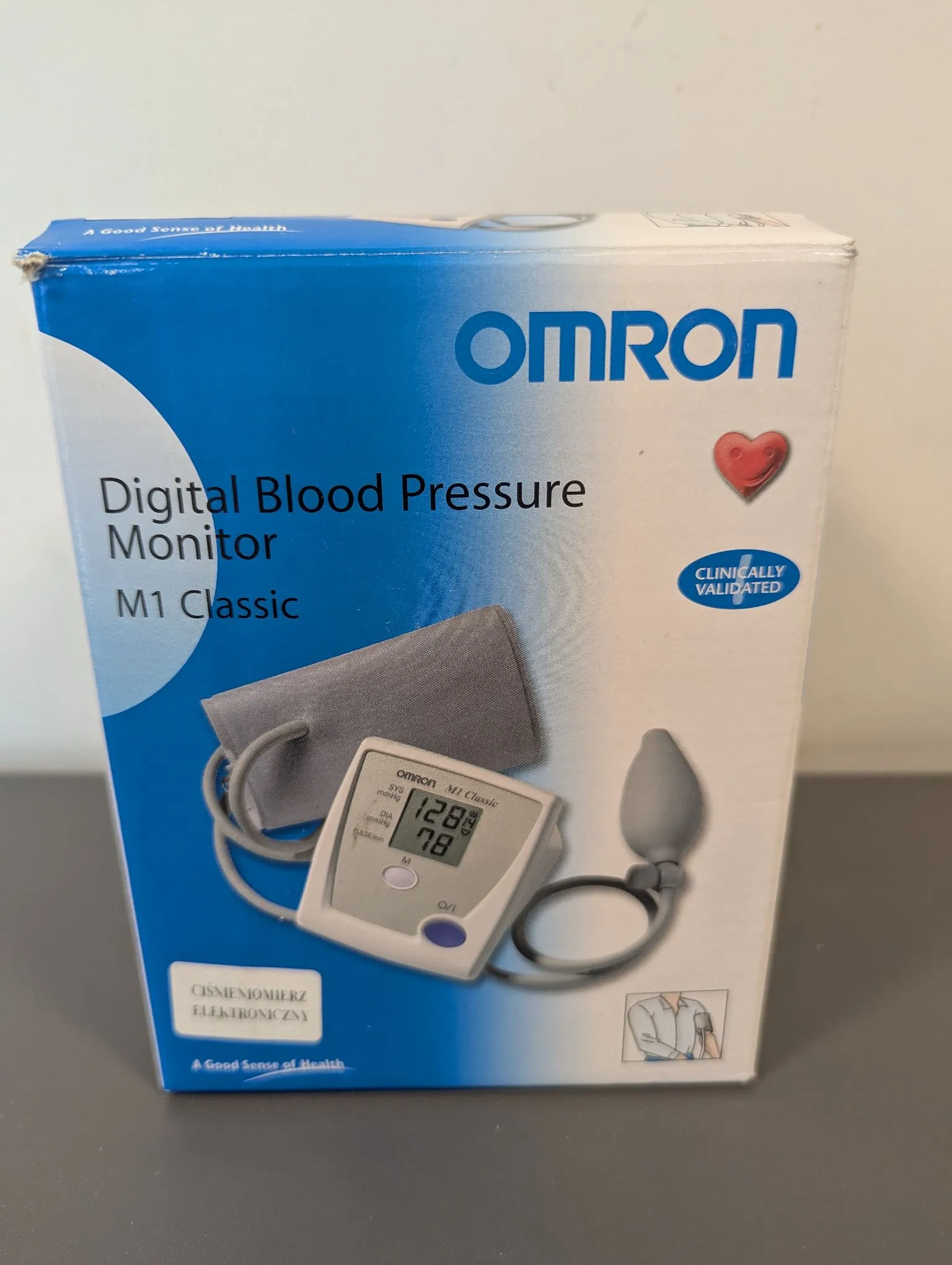 Kartonowe opakowanie cyfrowego ciśnieniomierza marki Omron, model M1 Classic. Na niebiesko-białym pudełku widnieje napis 'OMRON', 'Digital Blood Pressure Monitor M1 Classic' oraz logo czerwonego serca i napis 'CLINICALLY VALIDATED'. Na zdjęciu produktu widoczny jest aparat z wyświetlaczem pokazującym wyniki 128 i 78, mankiet oraz gruszka do pompowania. Na dole znajduje się polski napis 'CIŚNIENIOMIERZ ELEKTRONICZNY'.