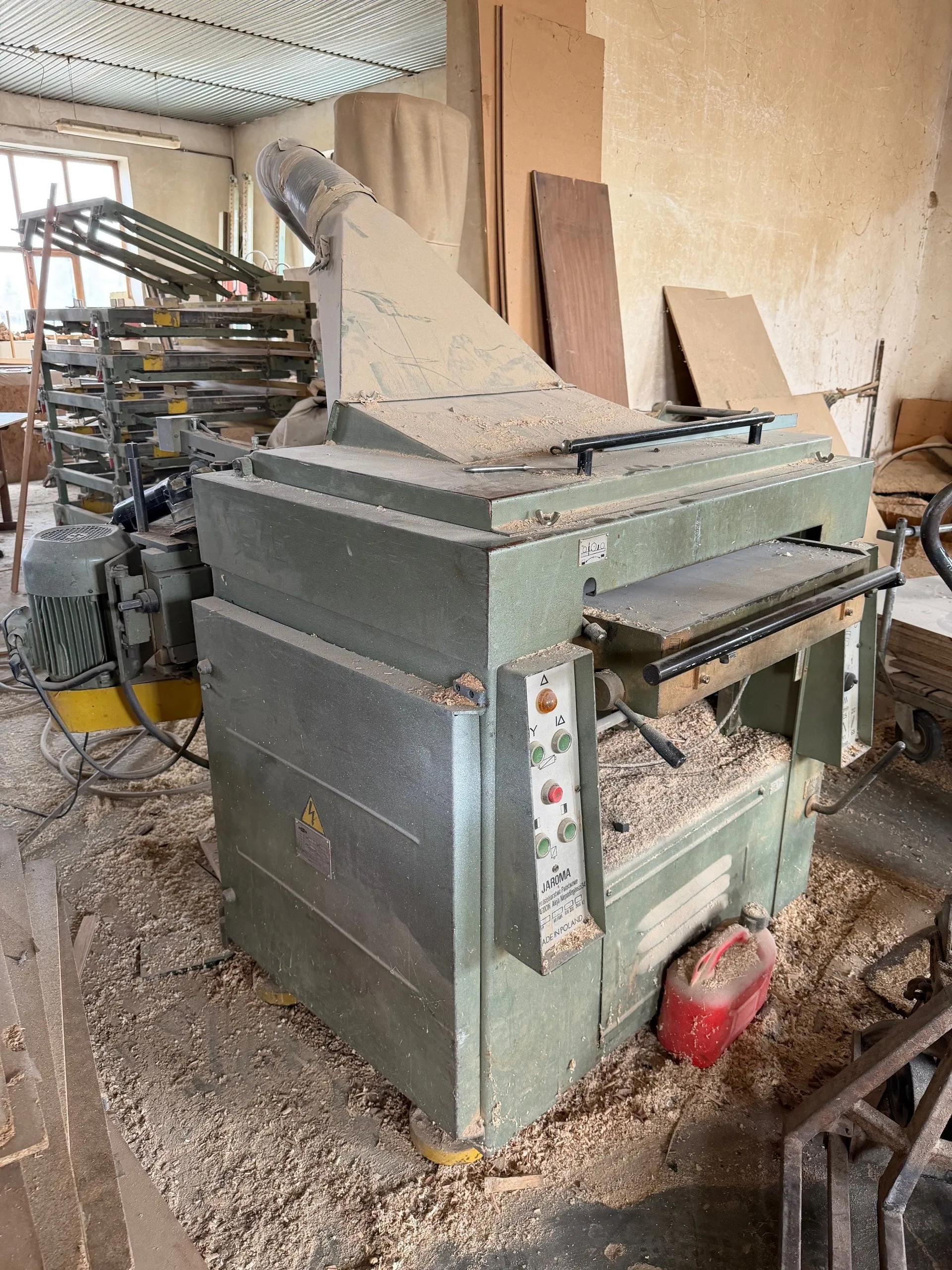 Wielki zielony maszynowy urządzenie przemysłowe w warsztacie, pokryte wiórami drewna. Na panelu sterowania widoczne przyciski oraz napis 'JAROMA'. Obok urządzenia czerwony pojemnik. W tle stosy desek, inne maszyny, okna i strop z blachy falistej. Ściany pomieszczenia są zużyte, a na maszynie umieszczony jest żółty znak ostrzegawczy z piktogramem przewodu elektrycznego.