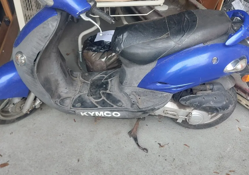Niebiesko-czarny skuter marki KYMCO w stanie zużycia, z uszkodzonym czarnym siedzeniem i widocznym napisem "KYMCO" na dolnej części. Skuter stoi na betonowej powierzchni, w tle widoczne są czarne worki na śmieci oraz fragmenty drewnianych i metalowych konstrukcji. Na podłodze leżą suche liście.