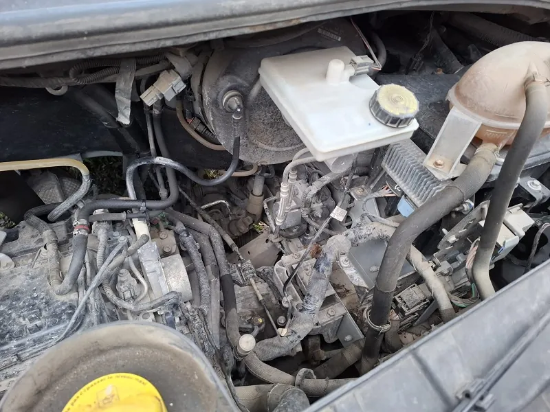 Zbliżenie na komorę silnika samochodu z widocznymi elementami takimi jak czarne przewody, rury, biały zbiornik z korkiem, żółty korek na dole po lewej stronie oraz zanieczyszczona powierzchnia silnika. Brak czytelnego tekstu.