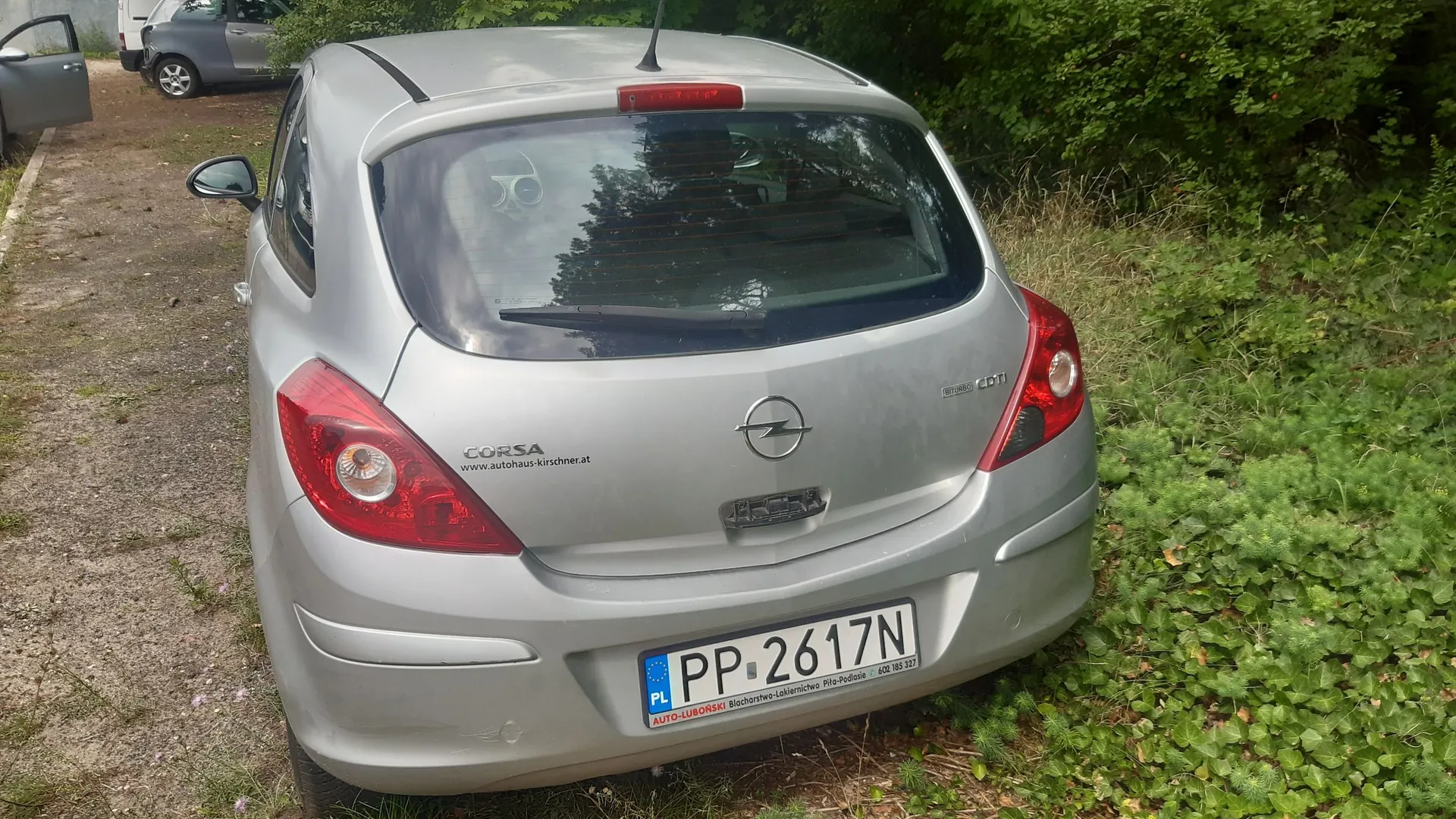 Srebrny samochód marki Opel Corsa zaparkowany na żwirowym podjeździe obok zielonych krzewów. Widoczny jest tył pojazdu z logo Opla, napisem CORSA oraz tablicą rejestracyjną o numerze PP 2617N. Na klapie bagażnika znajduje się również naklejka z adresem www.autohaus-kirschner.at oraz oznaczenie silnika CDTI.