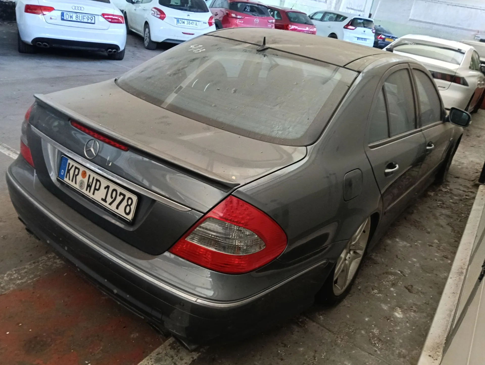 Szary samochód marki Mercedes-Benz zaparkowany na parkingu. Pojazd jest zakurzony, a na tylnej klapie widoczna jest tablica rejestracyjna o numerze KR WP 1978. W tle widoczne są inne zaparkowane samochody w kolorach białym i czerwonym.