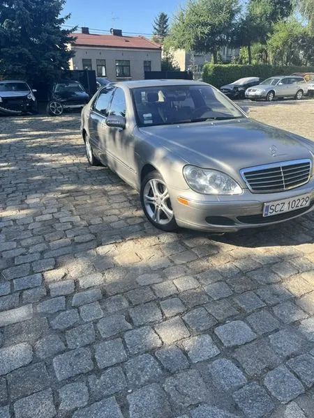 Srebrny samochód osobowy marki Mercedes-Benz zaparkowany na brukowanym placu. Auto jest ustawione pod kątem, widoczny jest przód i lewy bok pojazdu. Tablica rejestracyjna ma numer SCZ 10229. W tle widoczne są inne zaparkowane samochody, zielone drzewa oraz jasny budynek mieszkalny pod niebieskim niebem.